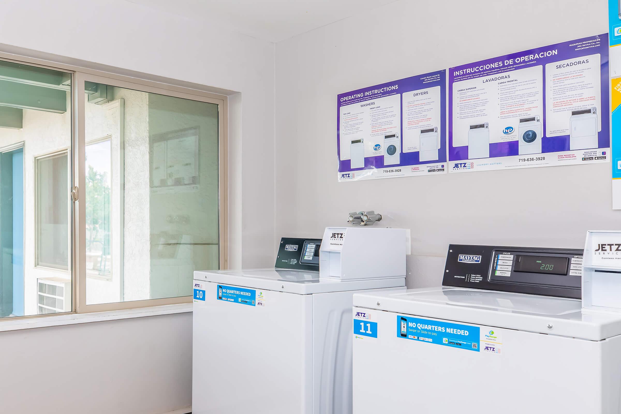 A clean laundry room featuring white washing machines and dryers. Instructional posters in English and Spanish are displayed on the wall above. A window allows natural light into the space, and the overall atmosphere is bright and organized.