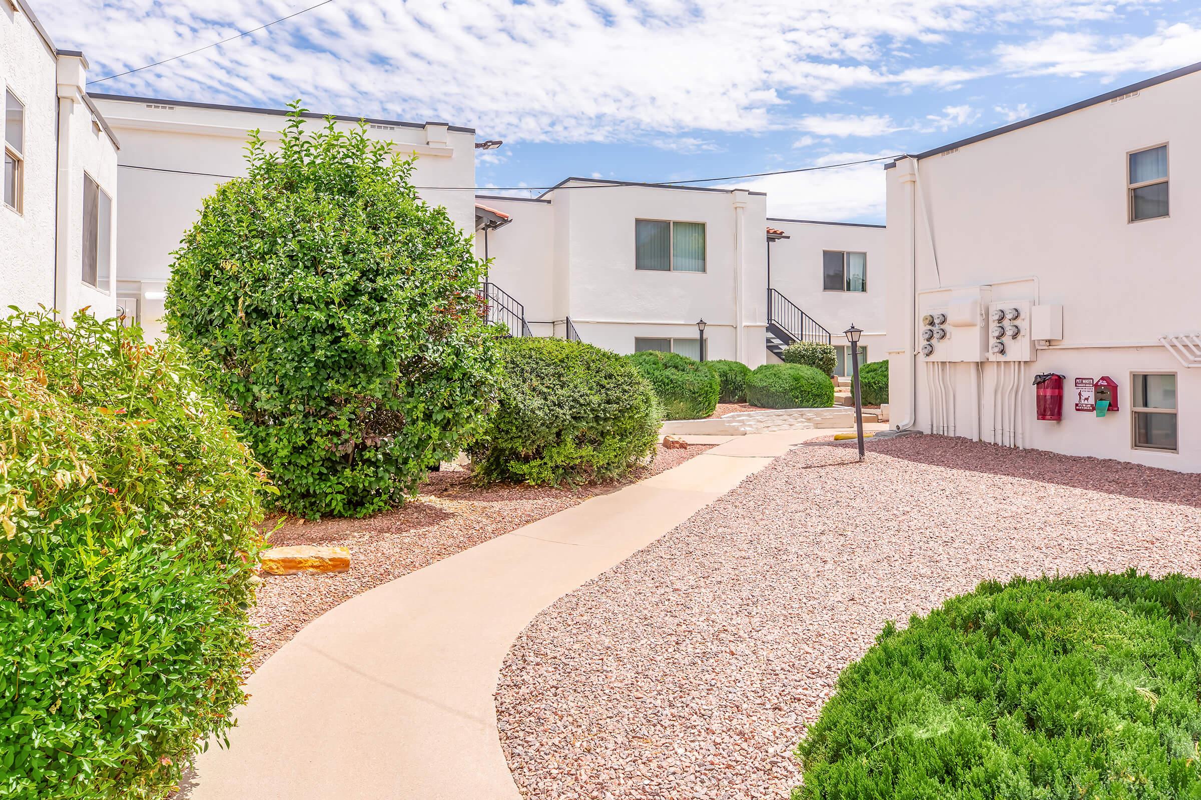 Lush green shrubs line a winding pathway through a landscaped area filled with gravel. White buildings with multiple windows are visible in the background, under a partly cloudy blue sky. The scene suggests a well-maintained residential or apartment complex.