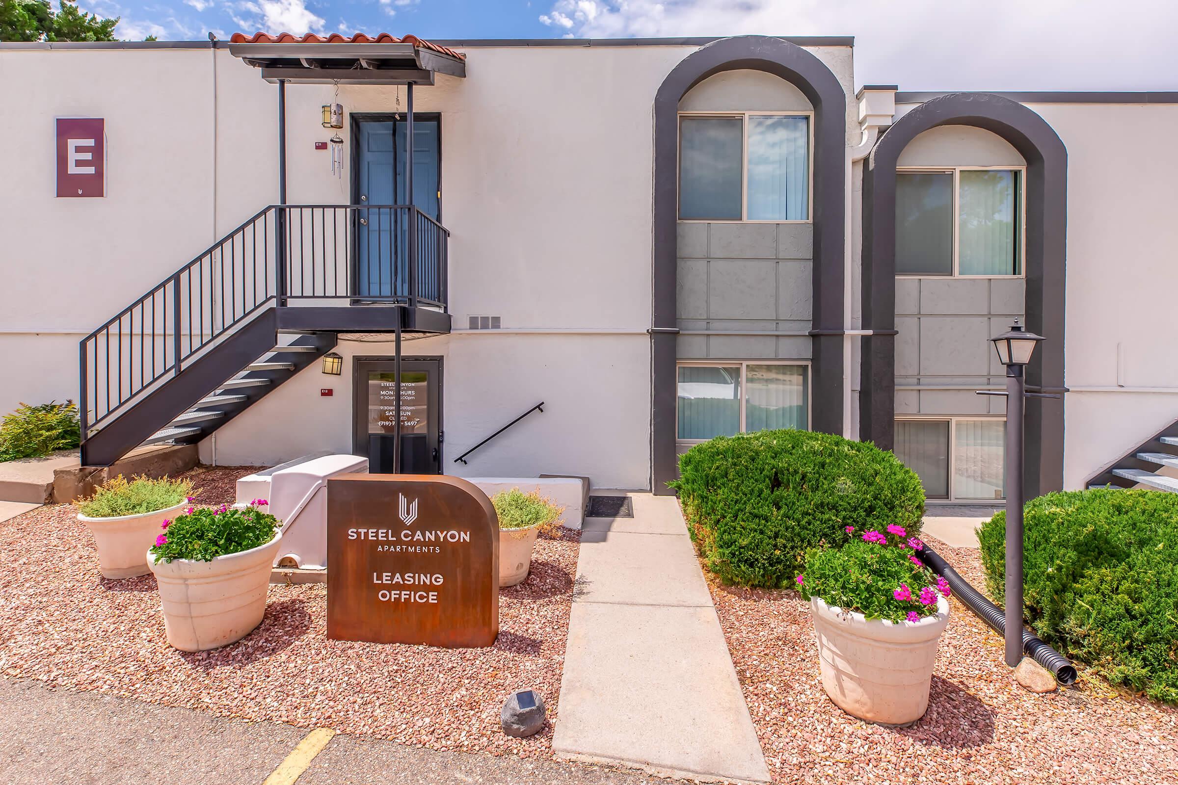 Leasing office entrance of Steel Canyon Apartments, featuring a modern building facade with a sign, stairs leading to the door, and well-maintained landscaping with flowering plants and shrubs. The atmosphere is bright, showcasing a welcoming entryway.