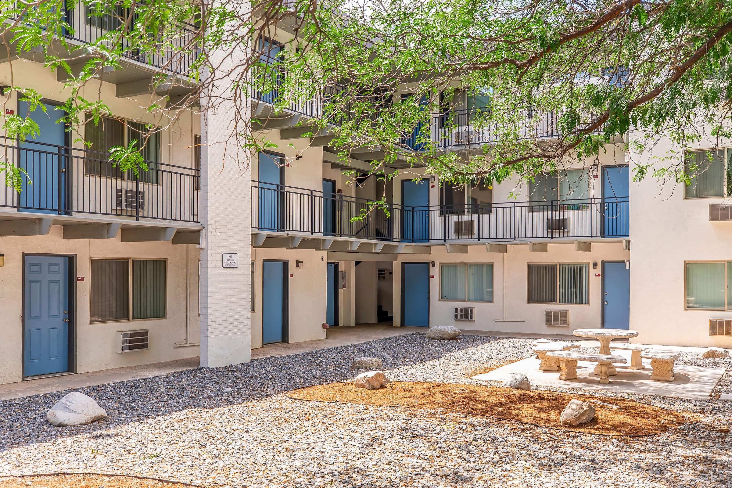 A view of a courtyard in an apartment complex featuring a gravel area, a stone table with benches, and several units with blue doors. Green trees provide shade, and the building has multiple levels with balconies. The overall atmosphere is inviting and relaxed.