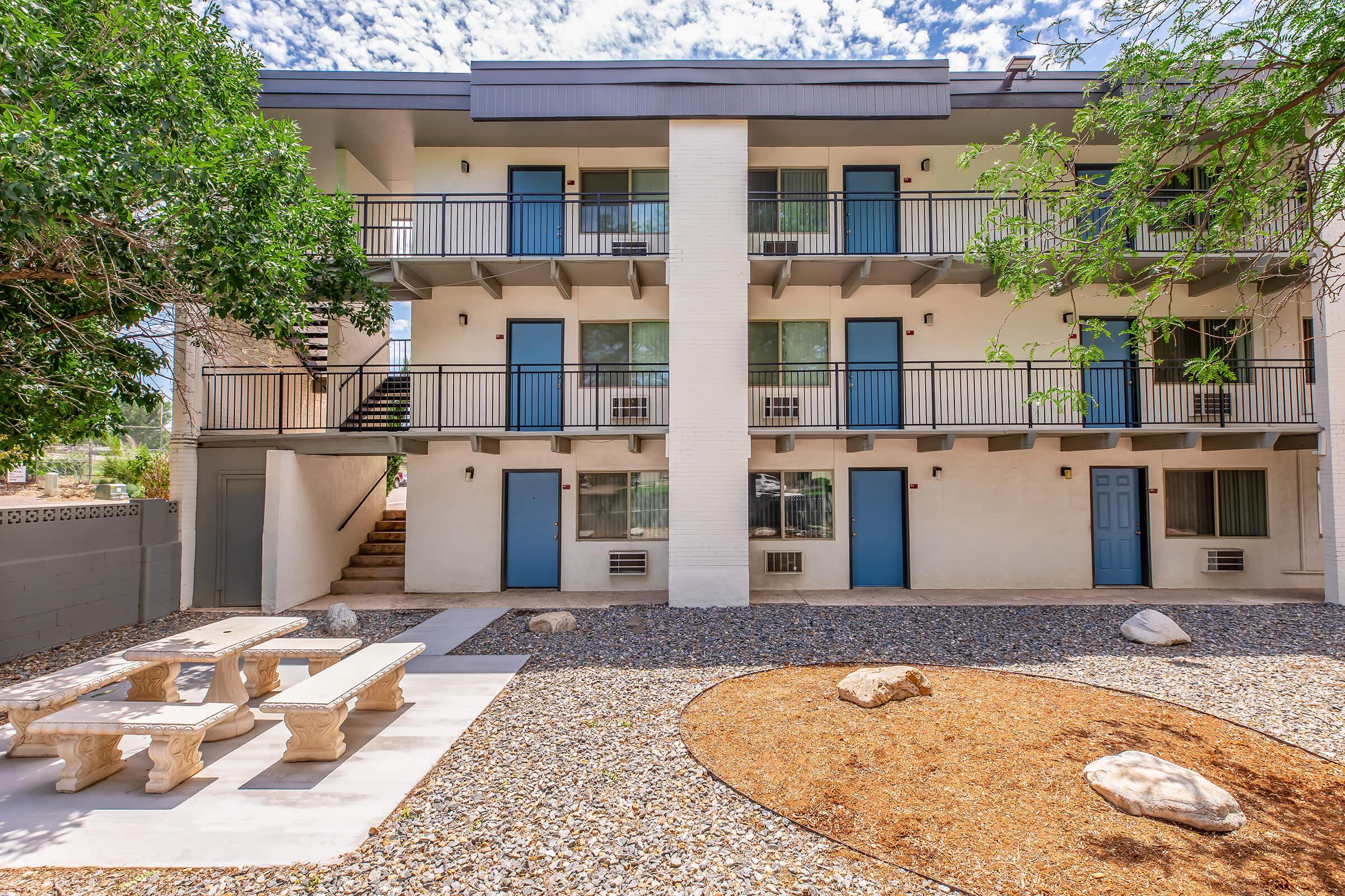 A view of a two-story building with multiple balconies. The building features blue doors and white walls, surrounded by a landscaped courtyard with gravel, a circular gravel area with rocks, and a picnic table. Lush green trees add shade to the outdoor space.