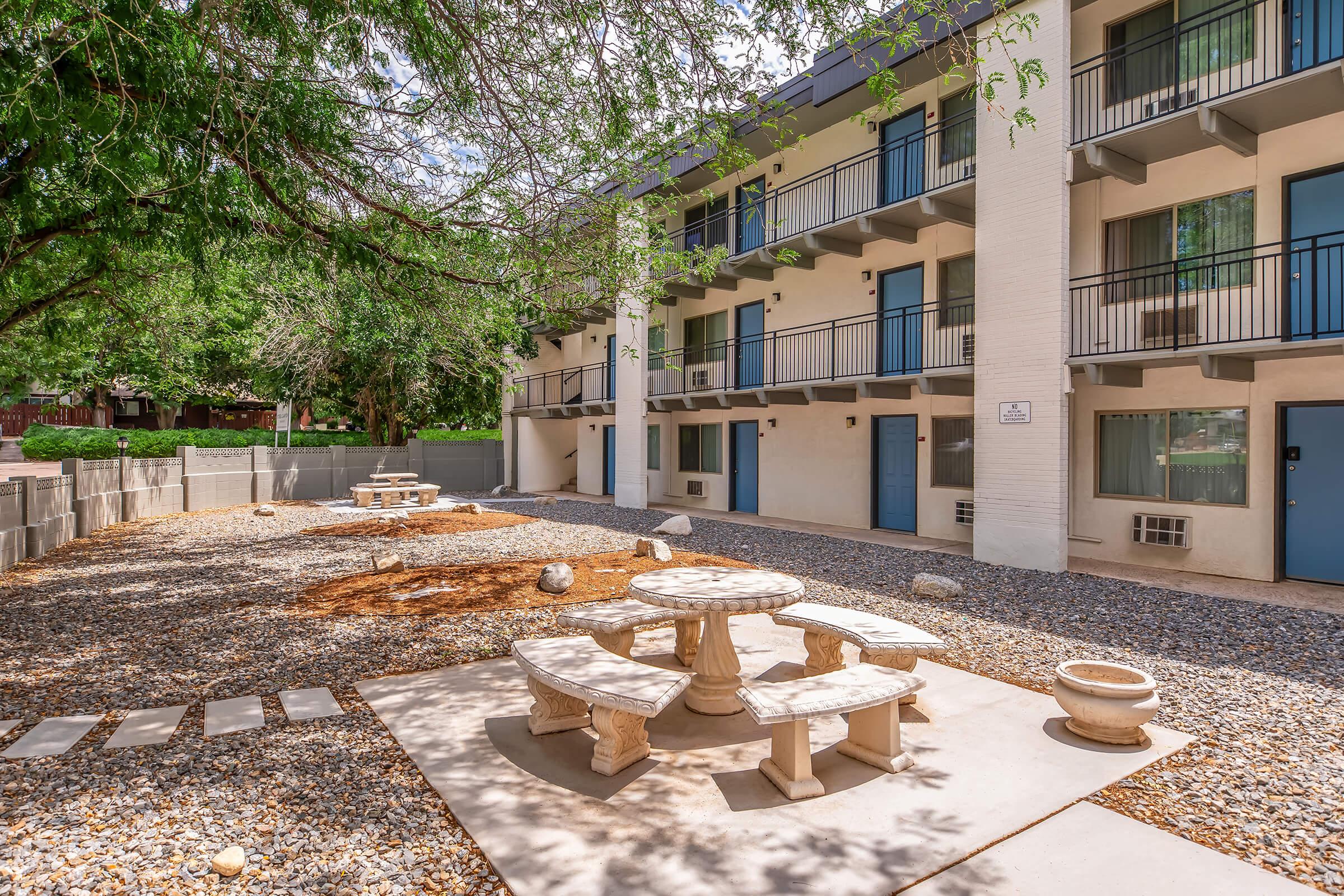 Outdoor seating area featuring a circular stone table surrounded by benches, situated in a gravel courtyard. In the background, a two-story building with balconies and blue doors is visible, along with greenery from surrounding trees. The scene is bright and inviting, ideal for relaxation.