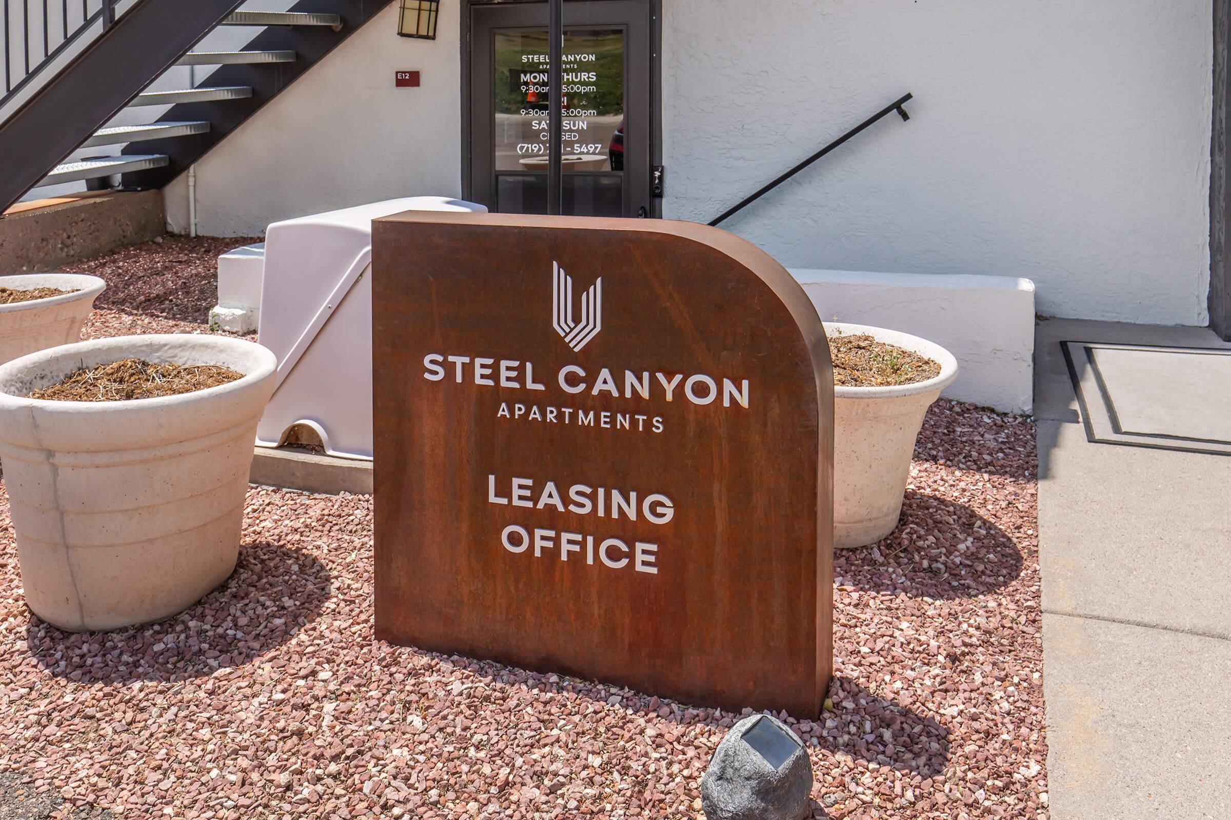 Sign for Steel Canyon Apartments Leasing Office, featuring a rust-colored metal design with white text. Surrounded by large planters filled with gravel or pebbles. The office entrance is visible in the background, framed by black stairs and a white wall.