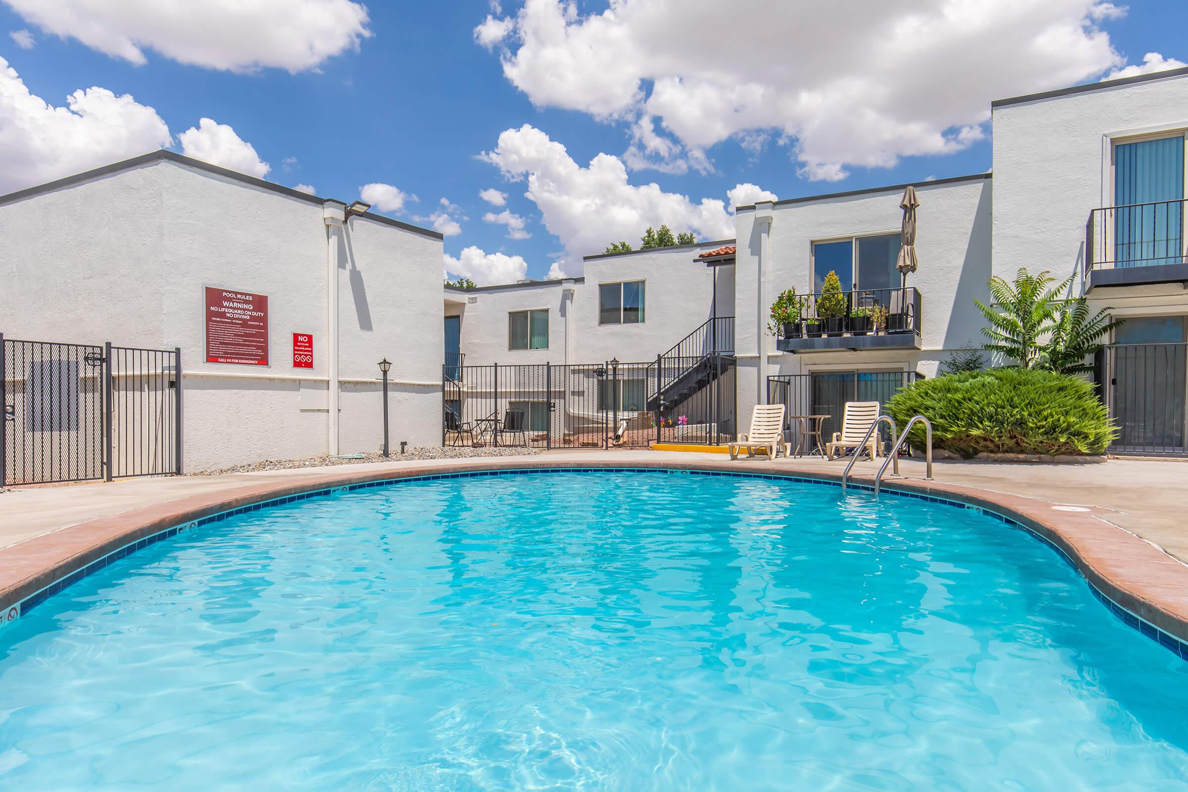 A clear blue swimming pool surrounded by lounge chairs, with white apartment buildings in the background. The scene is set under a bright sky filled with fluffy clouds, creating a relaxing atmosphere. There is a fenced area around the pool for privacy.
