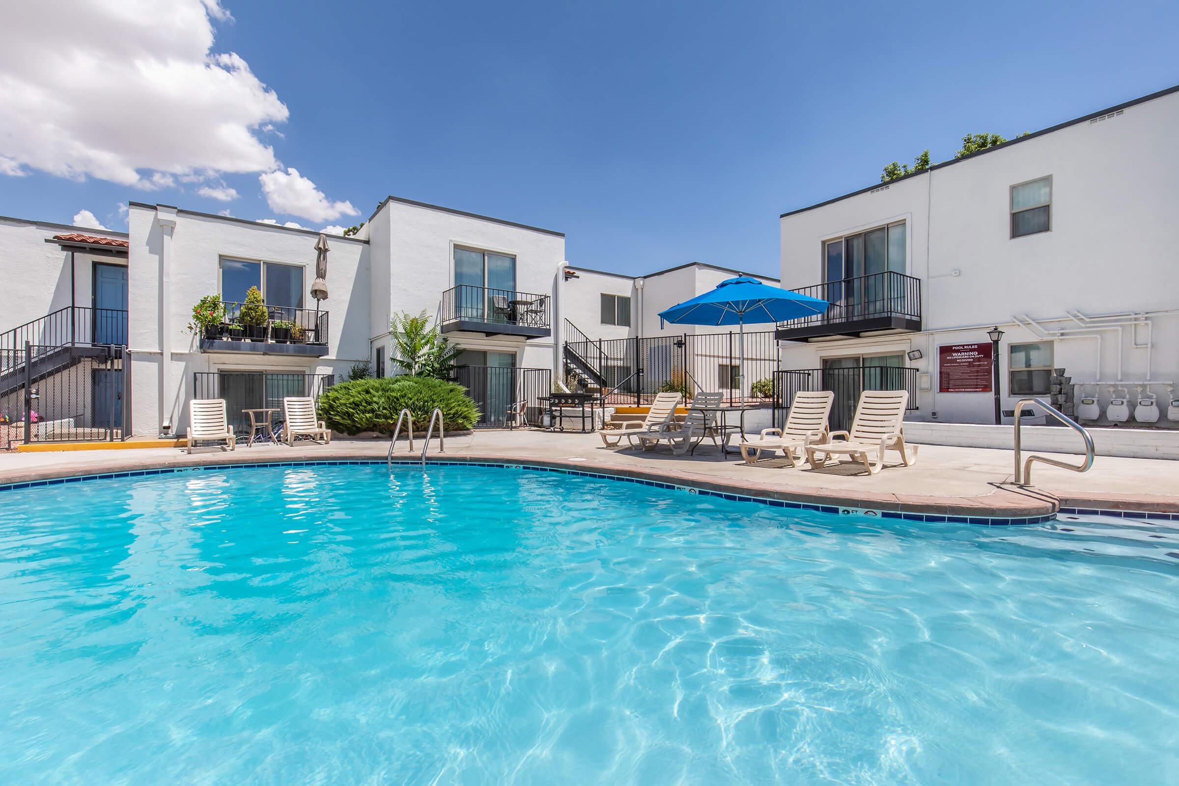 A clear blue swimming pool surrounded by lounge chairs and an umbrella, with white apartment buildings in the background. The sky is bright with a few clouds, creating a sunny and inviting atmosphere for relaxation and leisure.