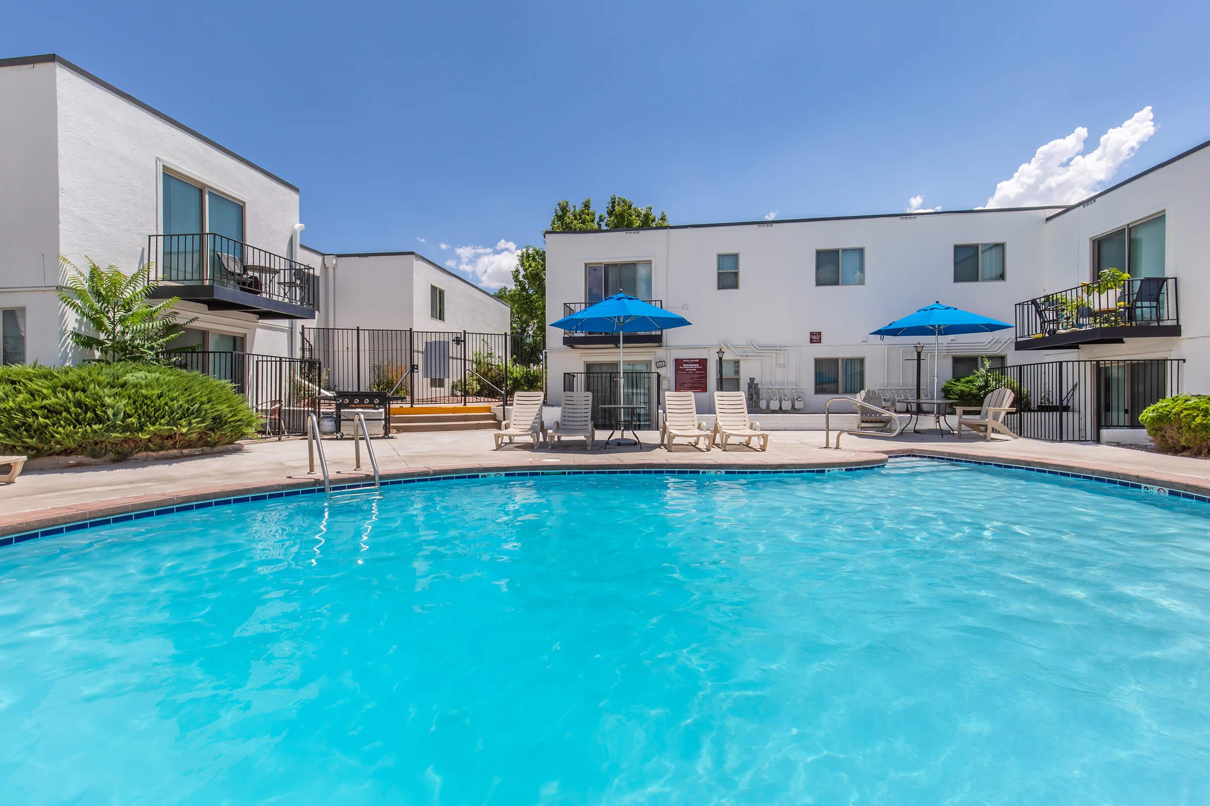 A clear swimming pool surrounded by lounge chairs and umbrellas, with white residential buildings in the background. Lush greenery lines the pool area, and the sky is bright blue with a few clouds scattered above.