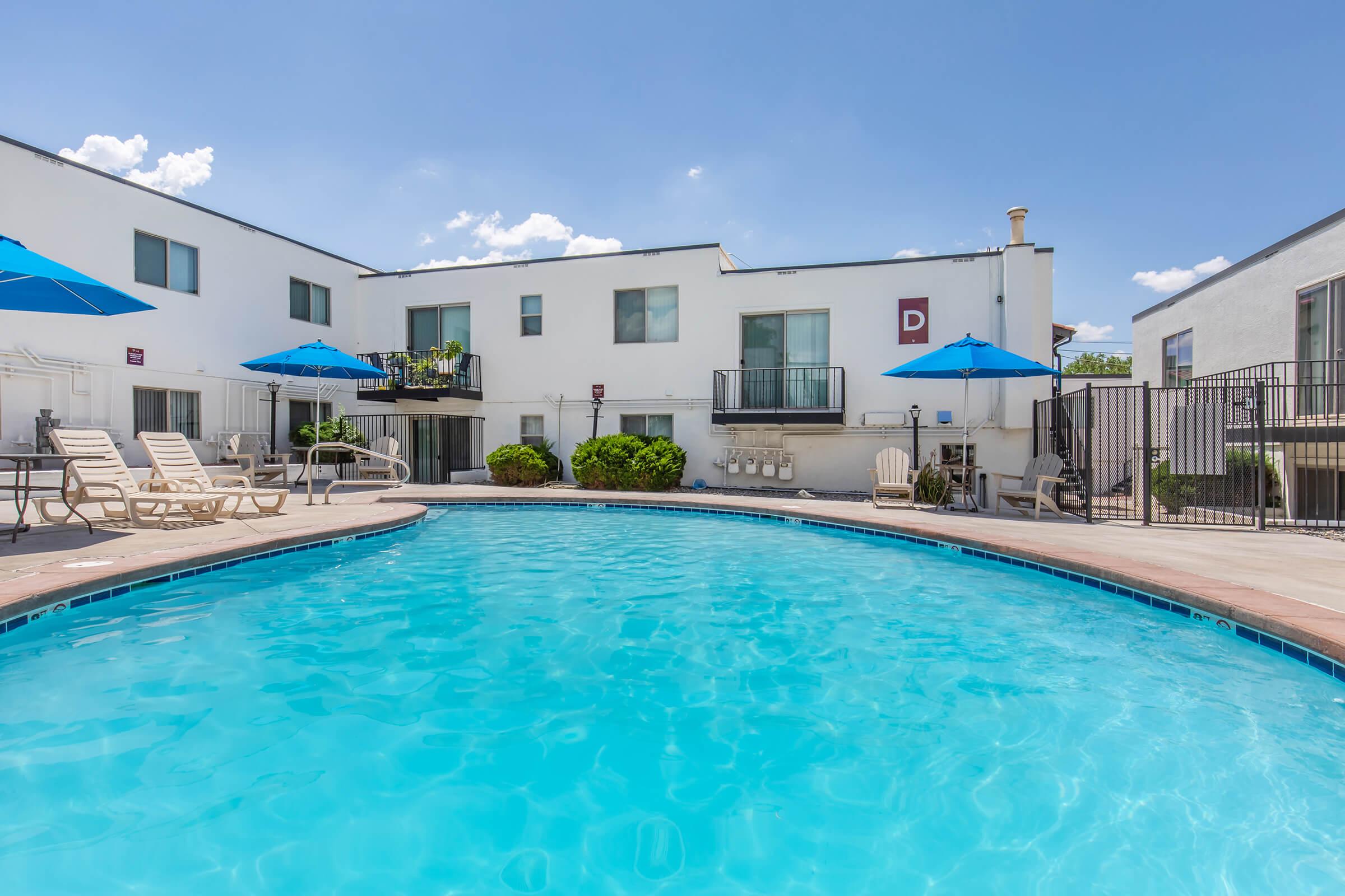 A bright outdoor swimming pool surrounded by lounge chairs and blue umbrellas. In the background, a white building features balconies and greenery. The sky is clear with a few clouds, creating a sunny, inviting atmosphere.
