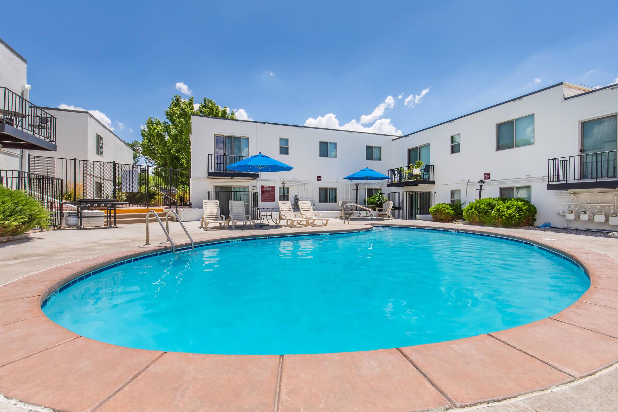 A clear blue swimming pool surrounded by a concrete deck. Lounge chairs with blue umbrellas are positioned nearby. In the background, modern white buildings are visible, with some greenery and a sunny sky adding to the inviting atmosphere.