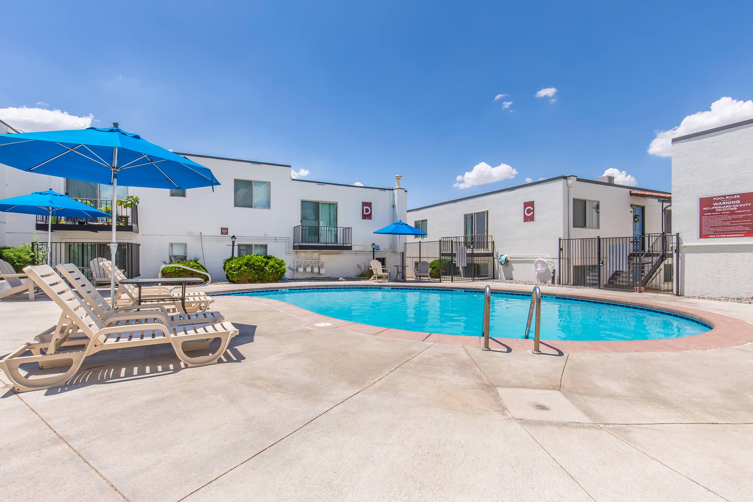 A swimming pool surrounded by lounge chairs and blue umbrellas, with two-story residential buildings in the background. The sky is clear and sunny, creating a bright and inviting atmosphere.
