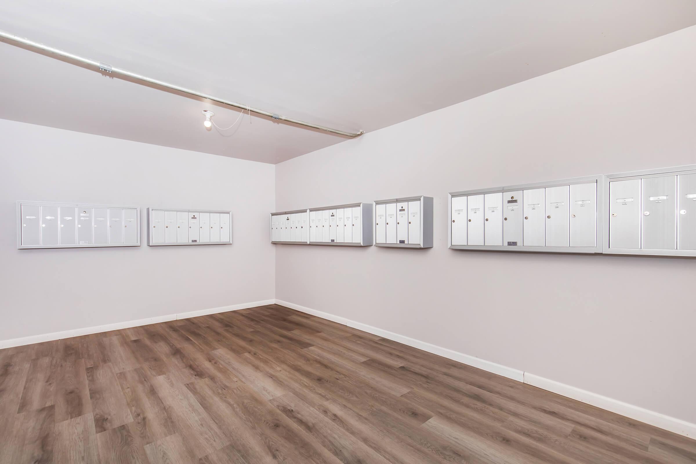 A bright, empty room with light-colored walls and wooden flooring, featuring several rows of white mailboxes mounted on the wall. The space is well-lit with overhead lighting and has a clean, minimalist appearance.