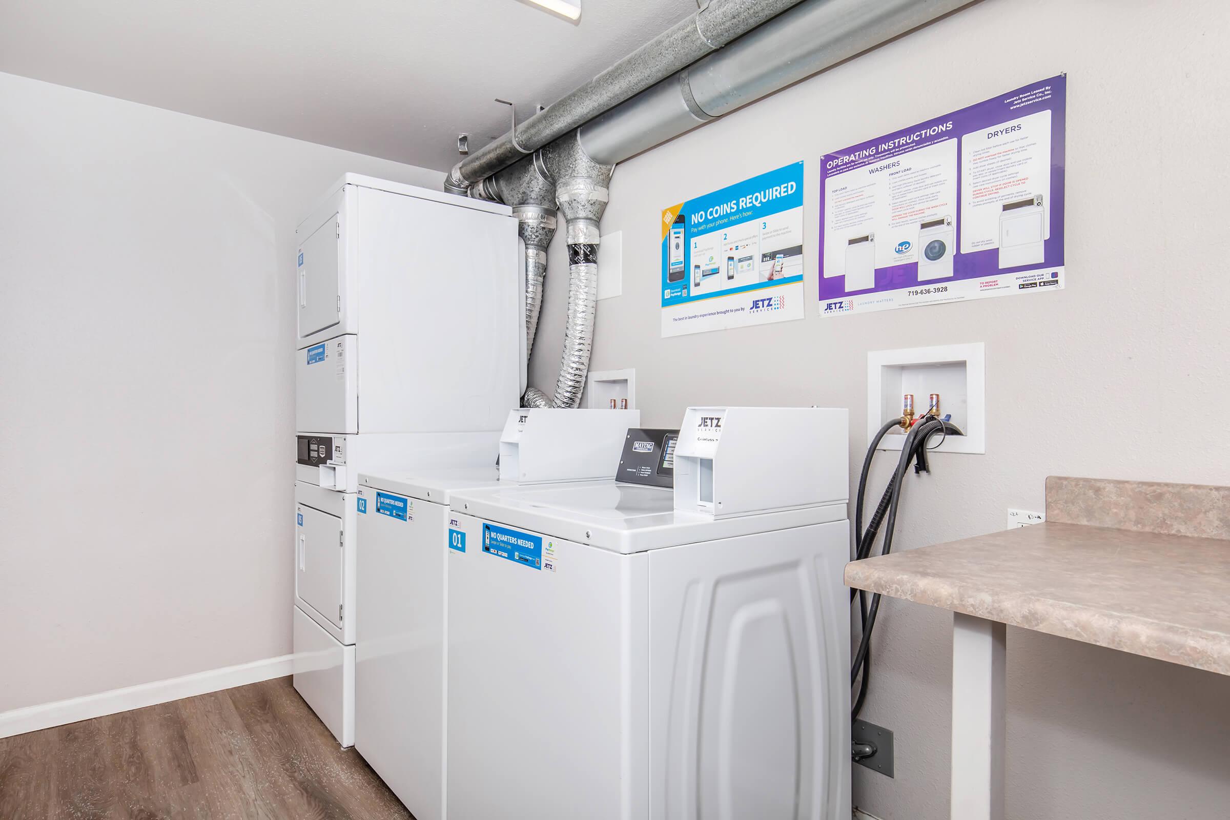 A small laundry room featuring a stacked washer and dryer unit, along with a front-loading washing machine. A countertop is visible next to the appliances. There are pipes and ventilation above, and instructional signage is attached to the wall nearby. The floor has laminate wood-like flooring.