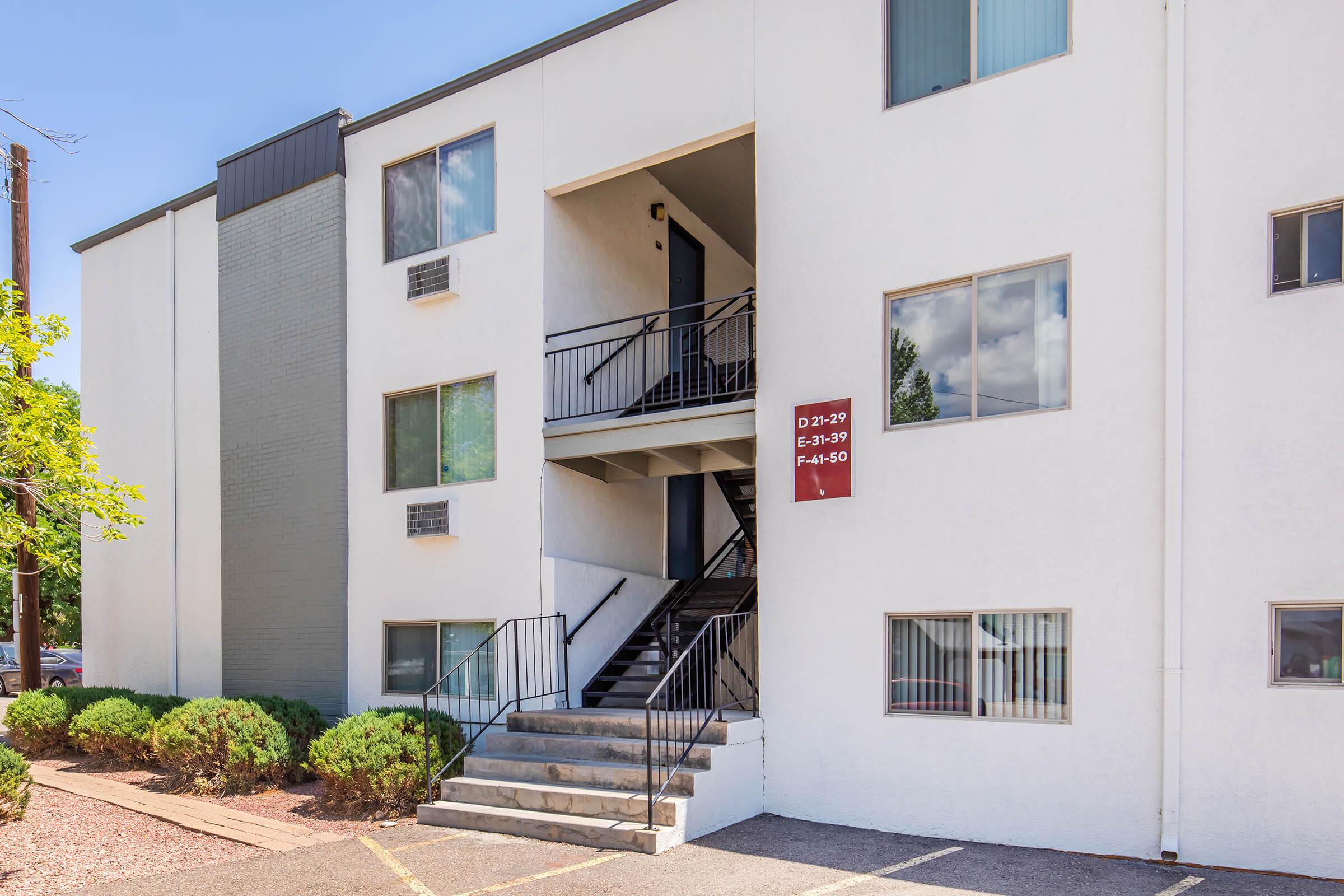 Exterior of a multistory apartment building featuring a staircase leading to a second-floor entry. The building has a light-colored facade with various windows, and a sign indicating apartment numbers near the entrance. Landscaping includes small shrubs around the base of the building. Bright blue sky in the background.