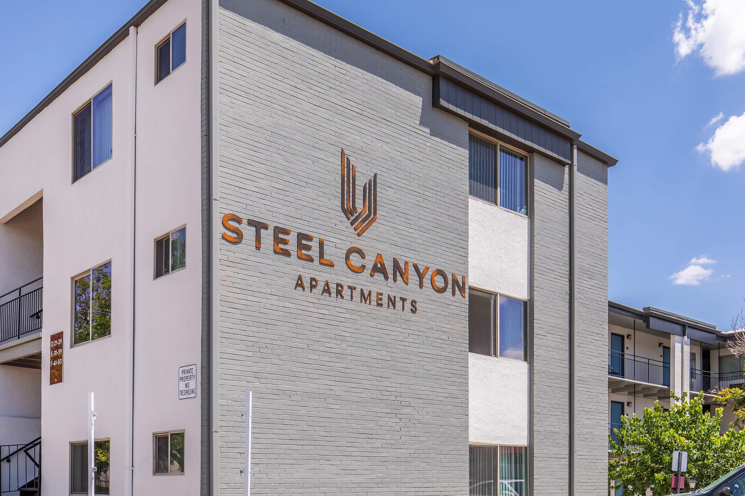 Exterior view of the Steel Canyon Apartments building, featuring a modern design with a gray brick facade. The prominent sign displays the name "STEEL CANYON" in large letters, with "APARTMENTS" beneath it. Windows are visible on multiple floors, surrounded by greenery. Blue sky with scattered clouds above.