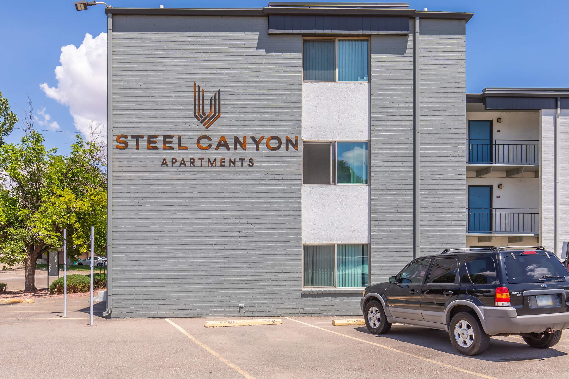 Exterior view of the Steel Canyon Apartments building, featuring a large sign with the name prominently displayed. The building is gray with multiple windows, and there is a black SUV parked in front. Lush greenery is visible nearby, under a clear blue sky with a few clouds.