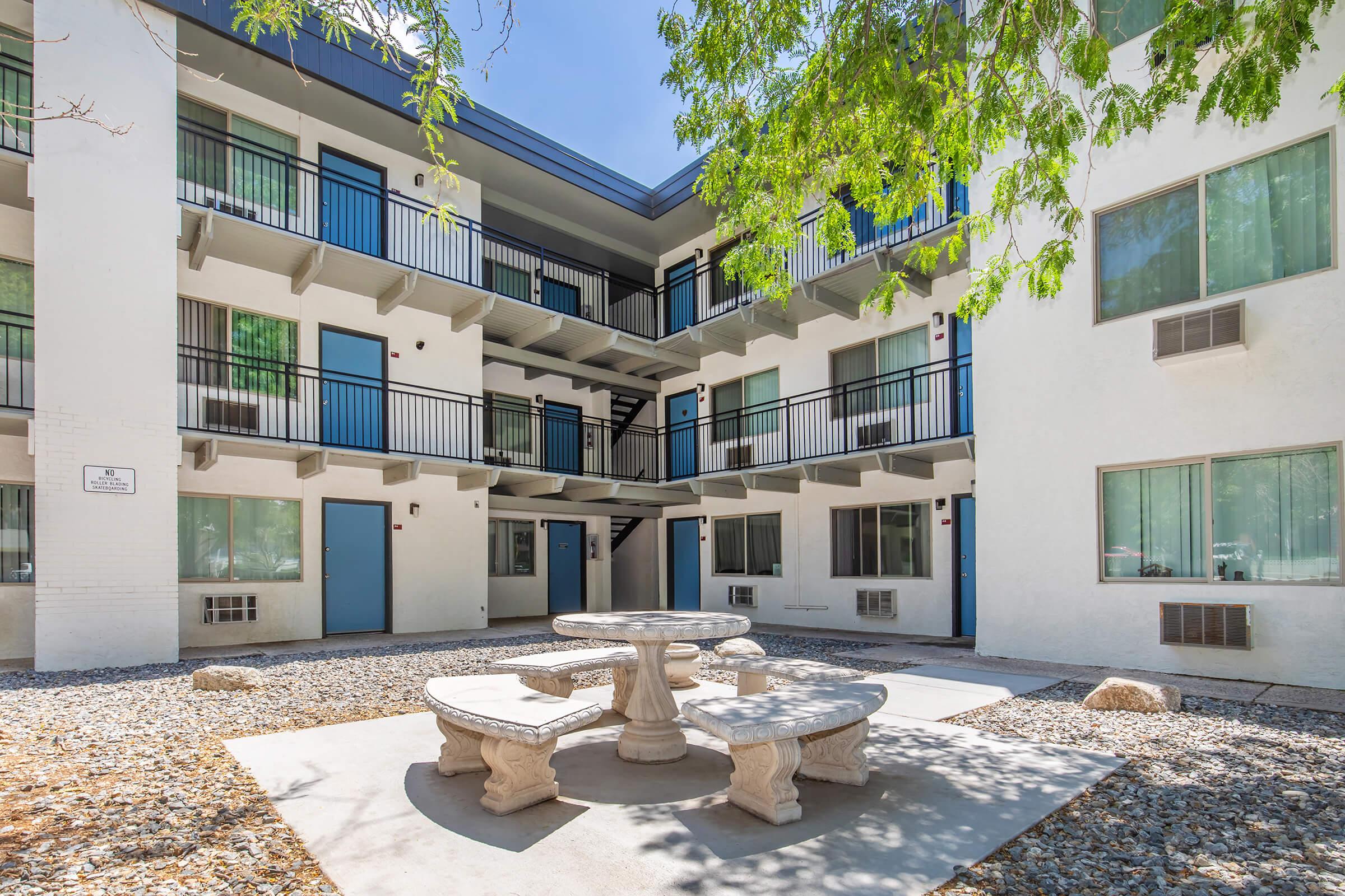 A view of a courtyard in an apartment complex, featuring two stories of white buildings with blue trim. The courtyard includes a stone table and benches surrounded by gravel and small rocks. There are several air conditioning units visible on the exterior walls, and greenery from nearby trees provides some shade.