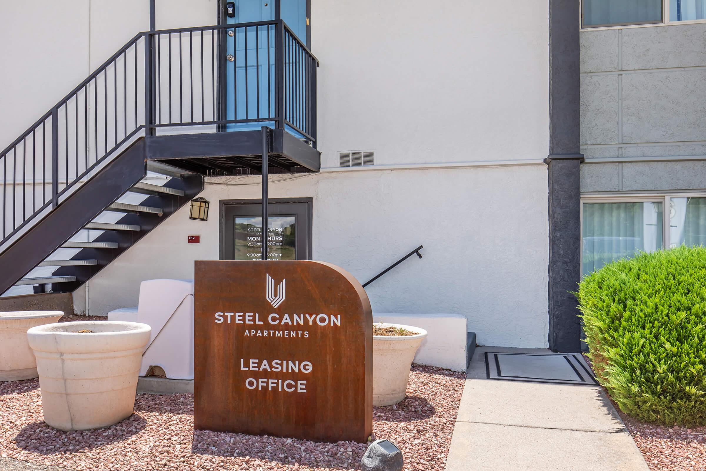 Sign for Steel Canyon Apartments with "LEASING OFFICE" prominently displayed, located at the entrance under a staircase. There are decorative plants in large pots, and a pathway leading to the entrance door. The building exterior features a mix of light and dark colors, with a clean, modern design.