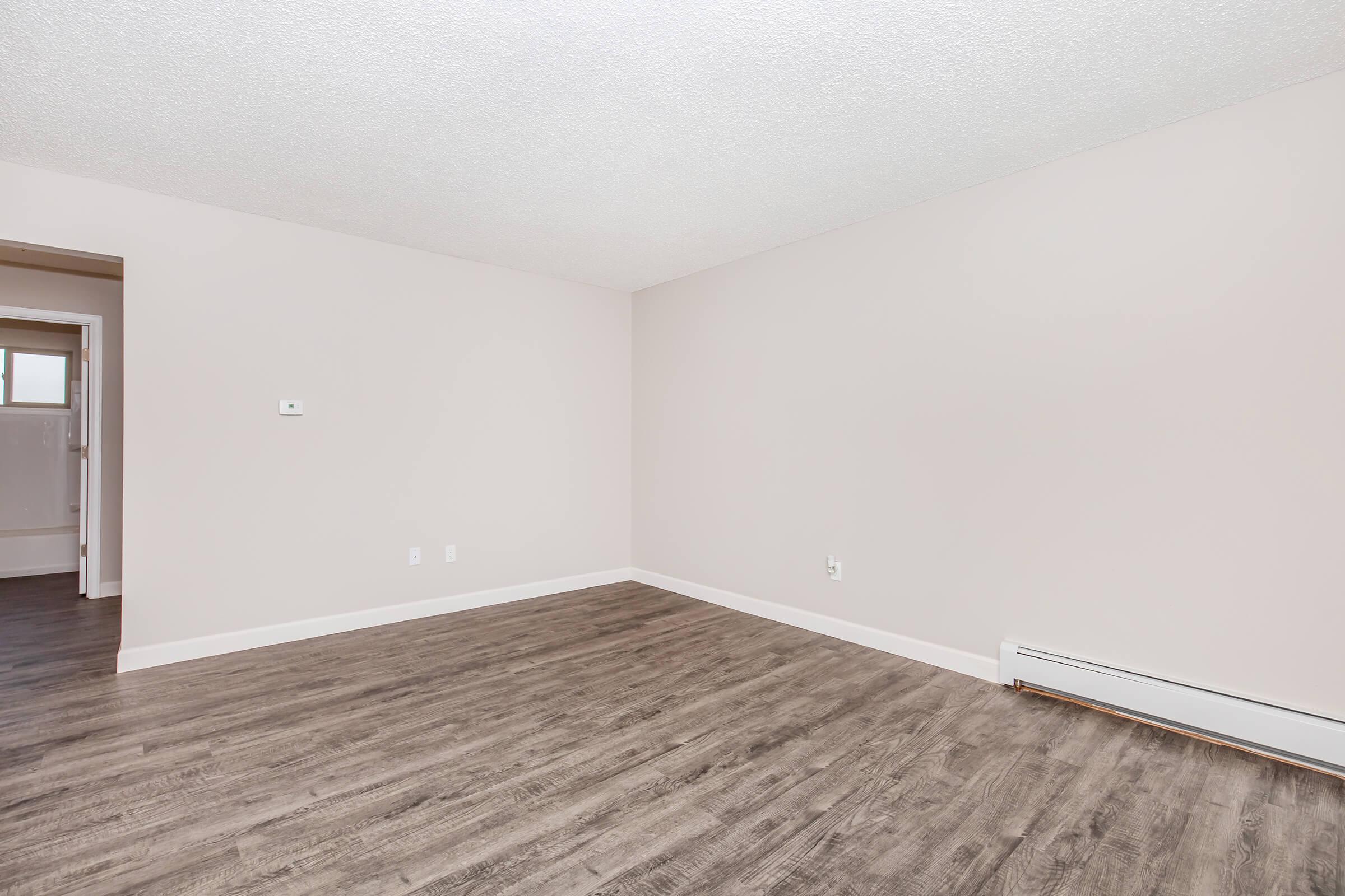 A spacious, light-colored room with a smooth, light brown wooden floor. The walls are painted in a neutral shade, and there are no furnishings. A doorway is visible at the left side, leading to another room. The overall atmosphere is clean and minimalistic, suitable for various uses.