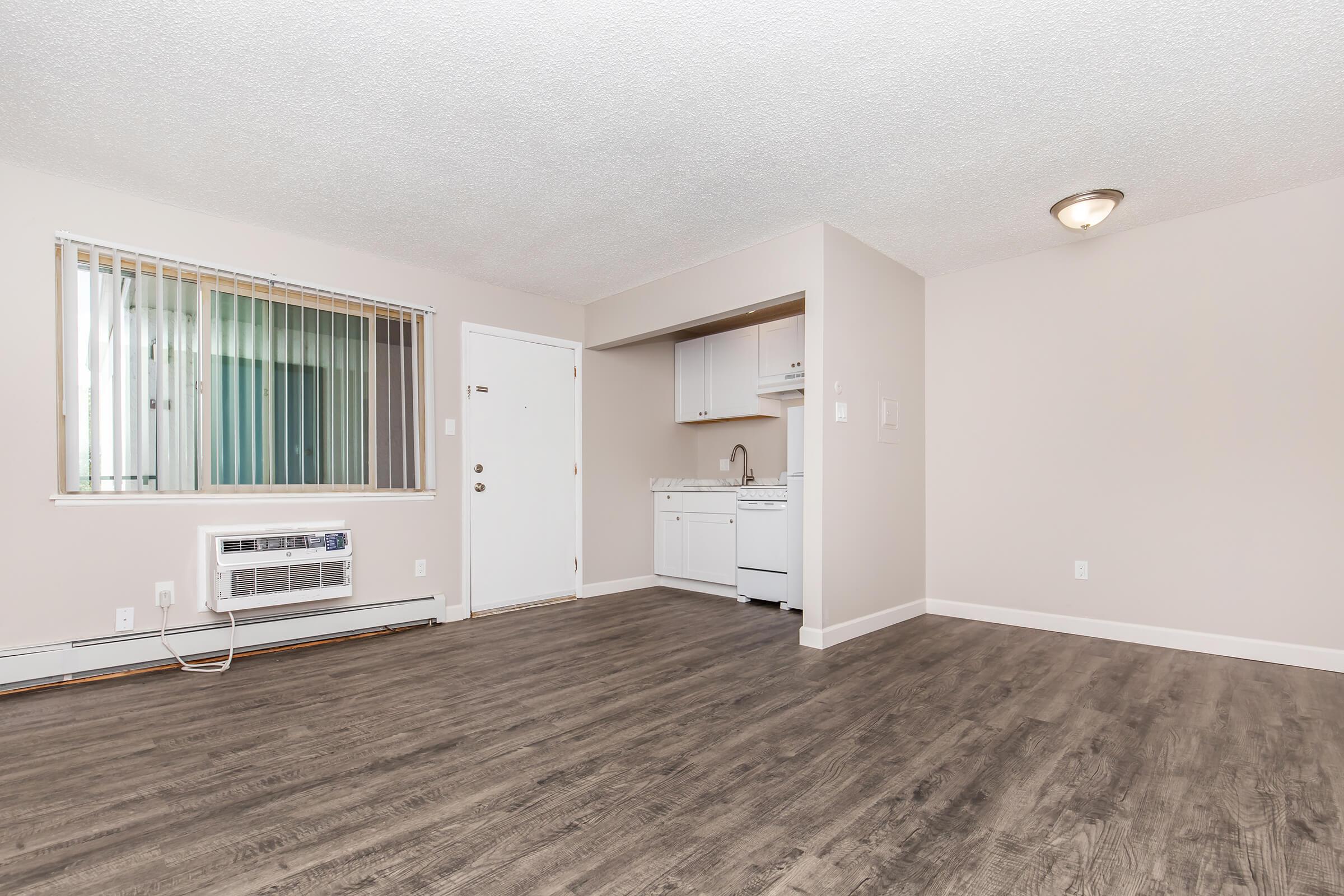 A view of a vacant apartment interior featuring light-colored walls, a wooden floor, and a kitchenette with a sink and cabinets. There's a door on the left, an air conditioning unit below the window, and ample natural light coming through the blinds. The ceiling is textured, and there’s a ceiling light fixture.