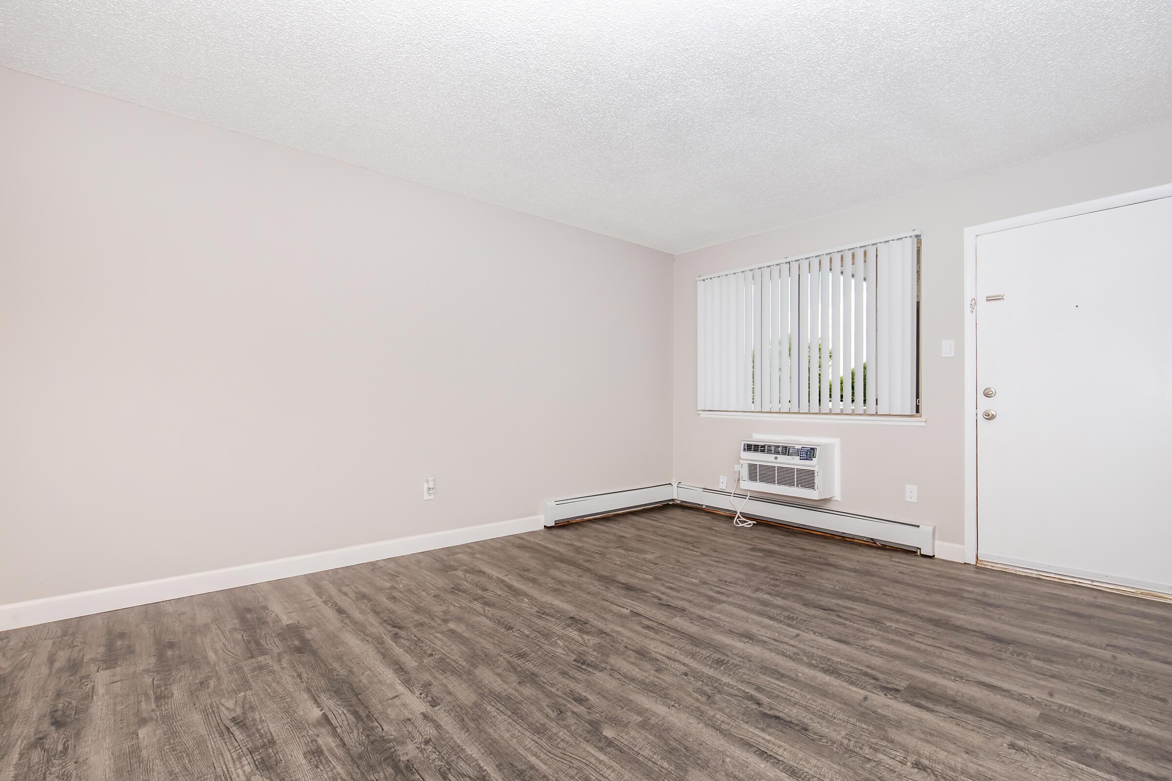 Empty room with light-colored walls, a window with vertical blinds, and an air conditioning unit on the floor. The flooring is a dark wood laminate, and there is a plain white door on the right side. The overall ambiance is clean and minimalistic, suitable for various uses.