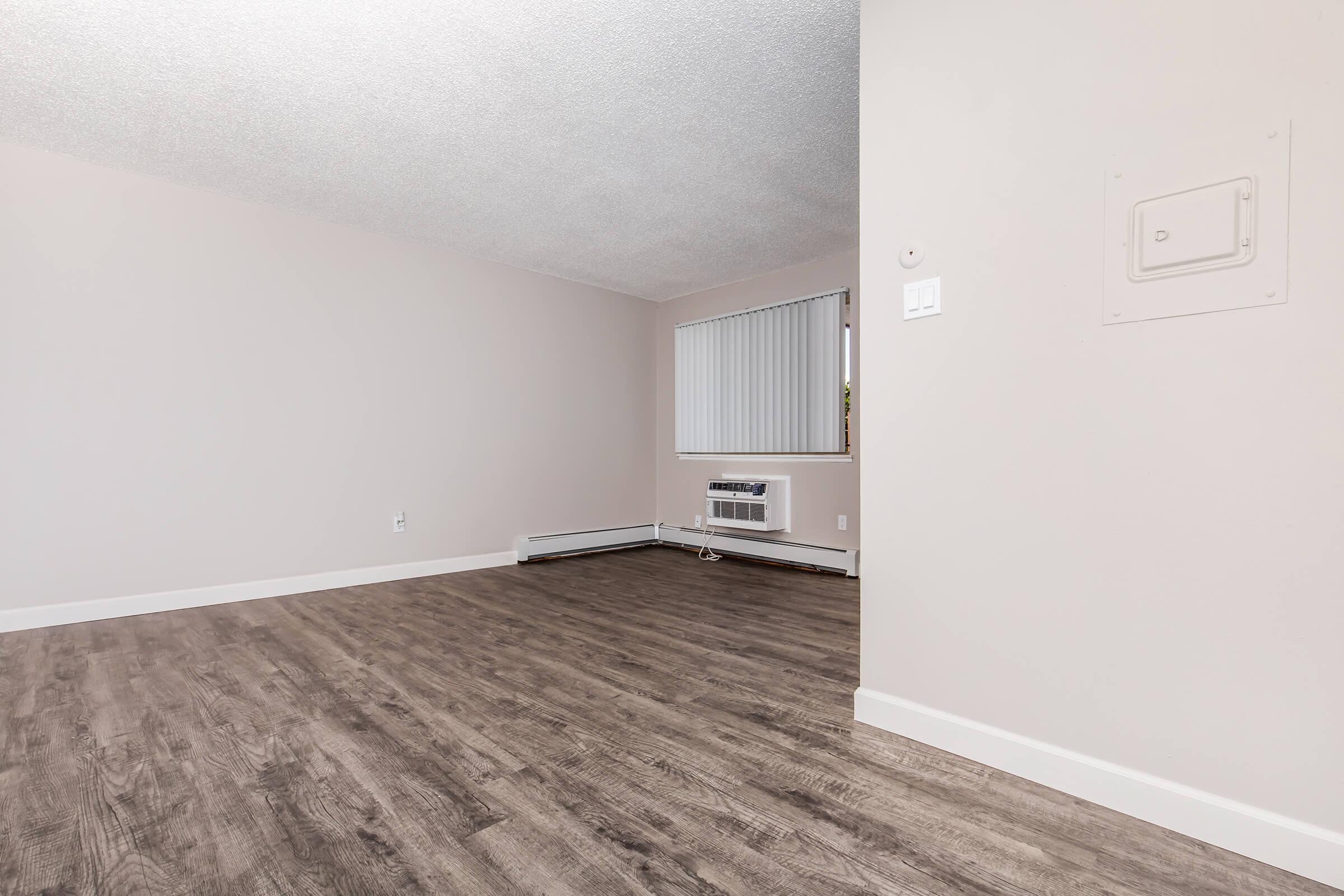 A spacious, empty room with light-colored walls and a textured ceiling. The floor is a wood-like laminate in a warm tone. There's an air conditioning unit beneath a window with vertical blinds, and the room features a baseboard heater. The lighting is bright and natural.