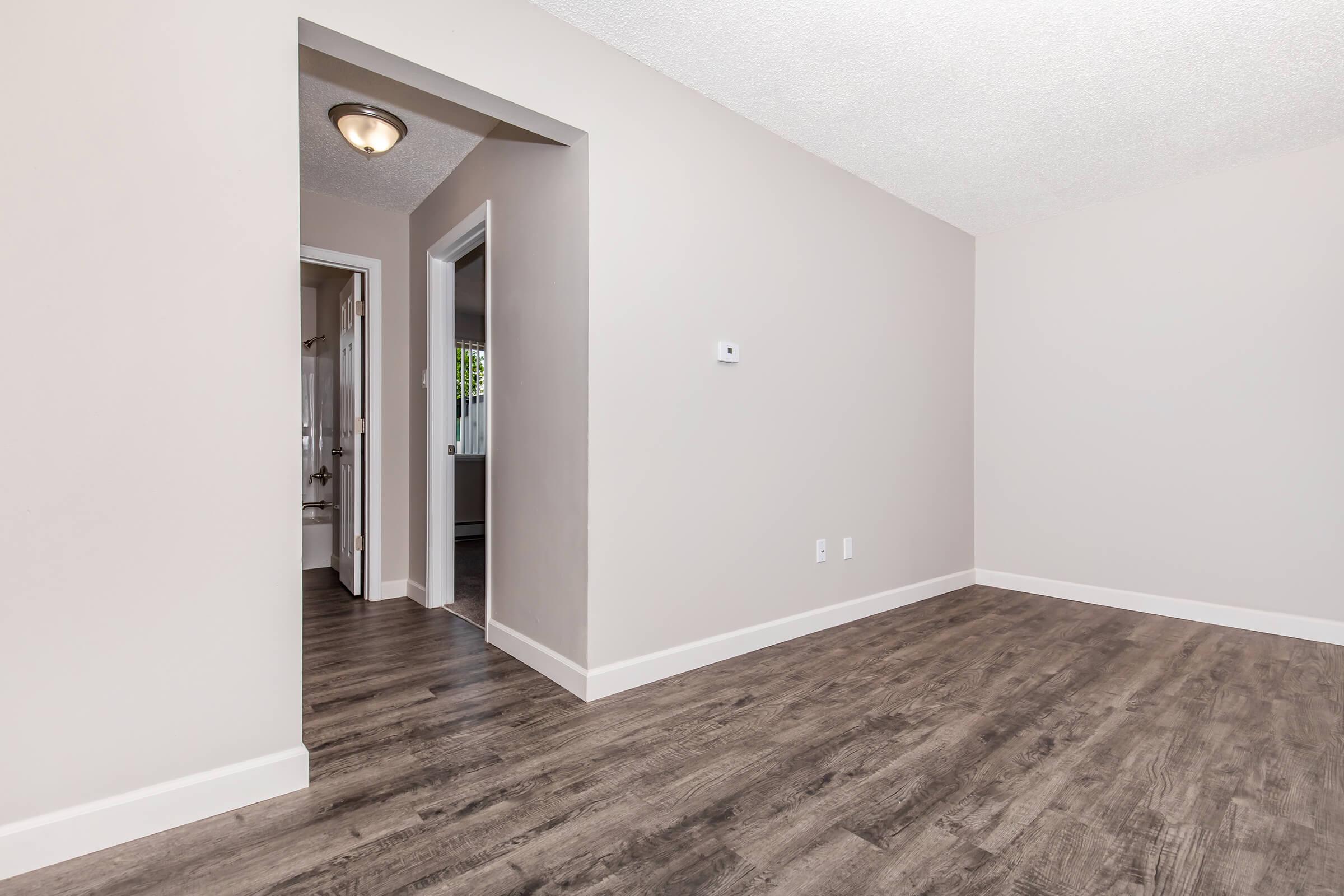 A spacious, light-filled room with light gray walls and new wood-like flooring. An archway leads to a hallway, while a door in the distance opens to another space. The area is uncluttered, ideal for various furniture arrangements. Natural light brightens the interior, creating an inviting atmosphere.