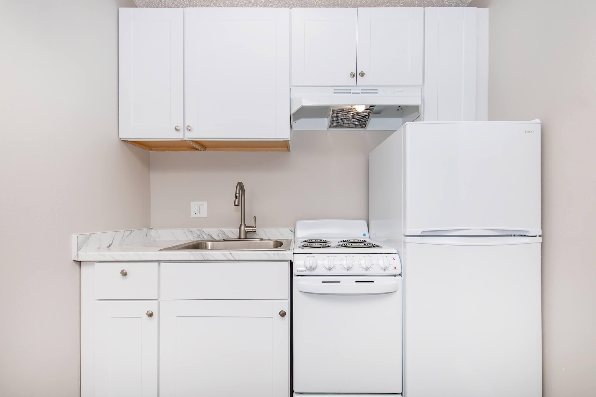 Compact kitchen featuring white cabinetry, a stainless steel sink, a stovetop with oven, and a refrigerator. The countertops are light-colored marble, and the walls are painted a neutral shade, creating a clean and modern aesthetic.