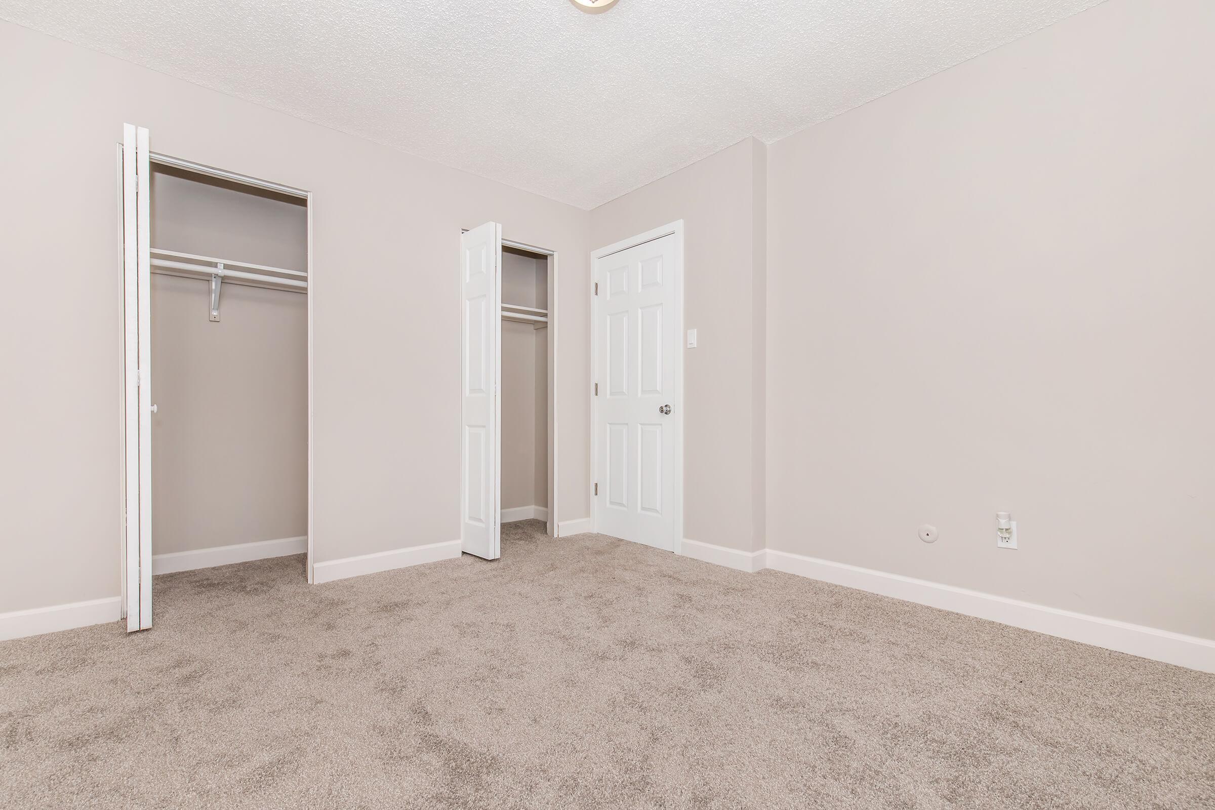A spacious, empty room with light beige carpet and soft, neutral-colored walls. There are two open doors: one leading to a closet on the left and the other to a standard door on the right. The room is well-lit, featuring a ceiling light fixture, and has a clean, minimalist aesthetic.
