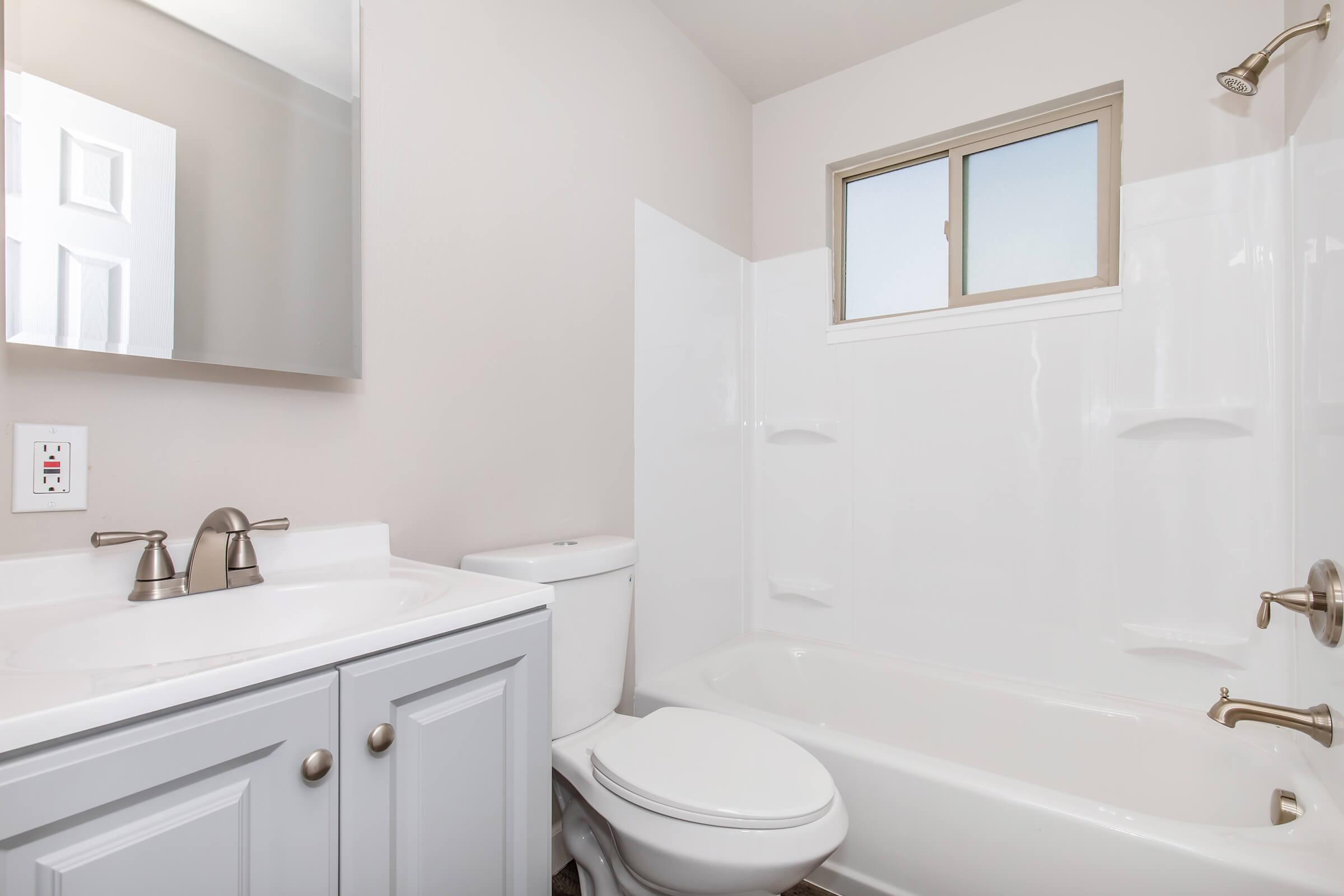 A clean, modern bathroom featuring a white bathtub and shower combination, a white toilet, and a vanity with a sink and mirror. The walls are painted a soft beige, and there is a window allowing natural light to enter the space.