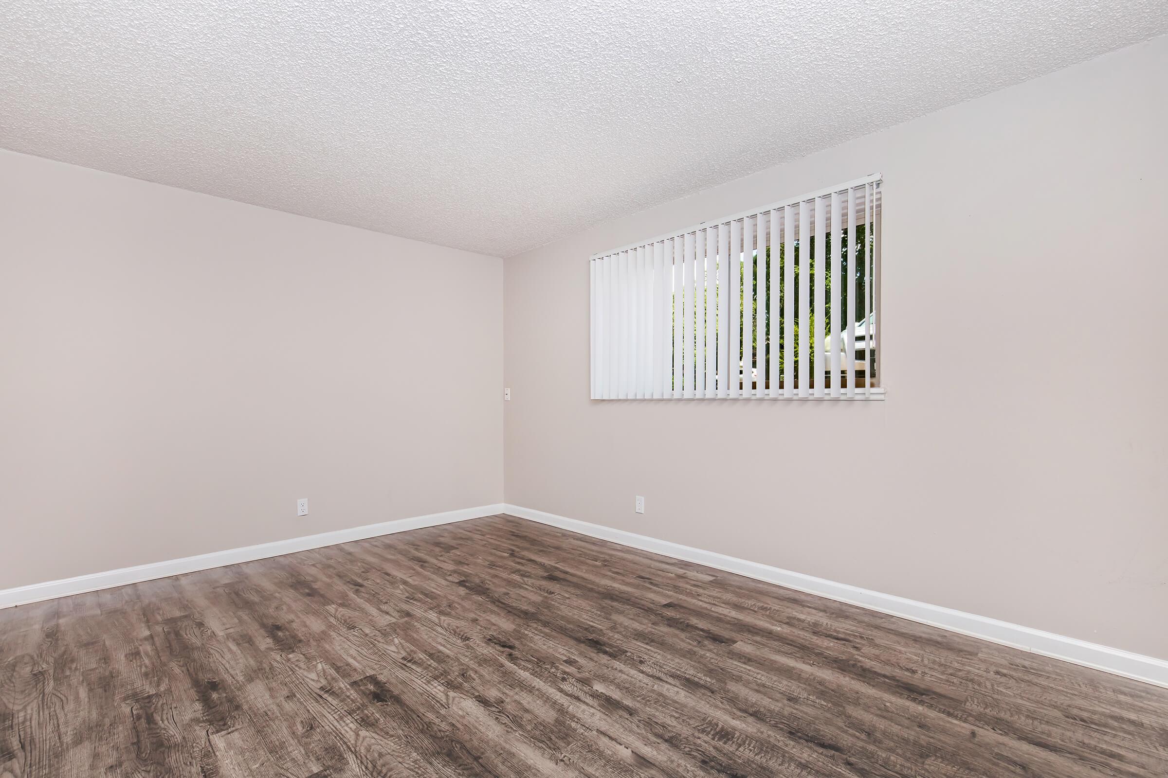 Empty room with light tan walls and a smooth wood-like floor. A window with white vertical blinds allows natural light to enter. The room features minimal decor and is spacious, creating a clean, open atmosphere.