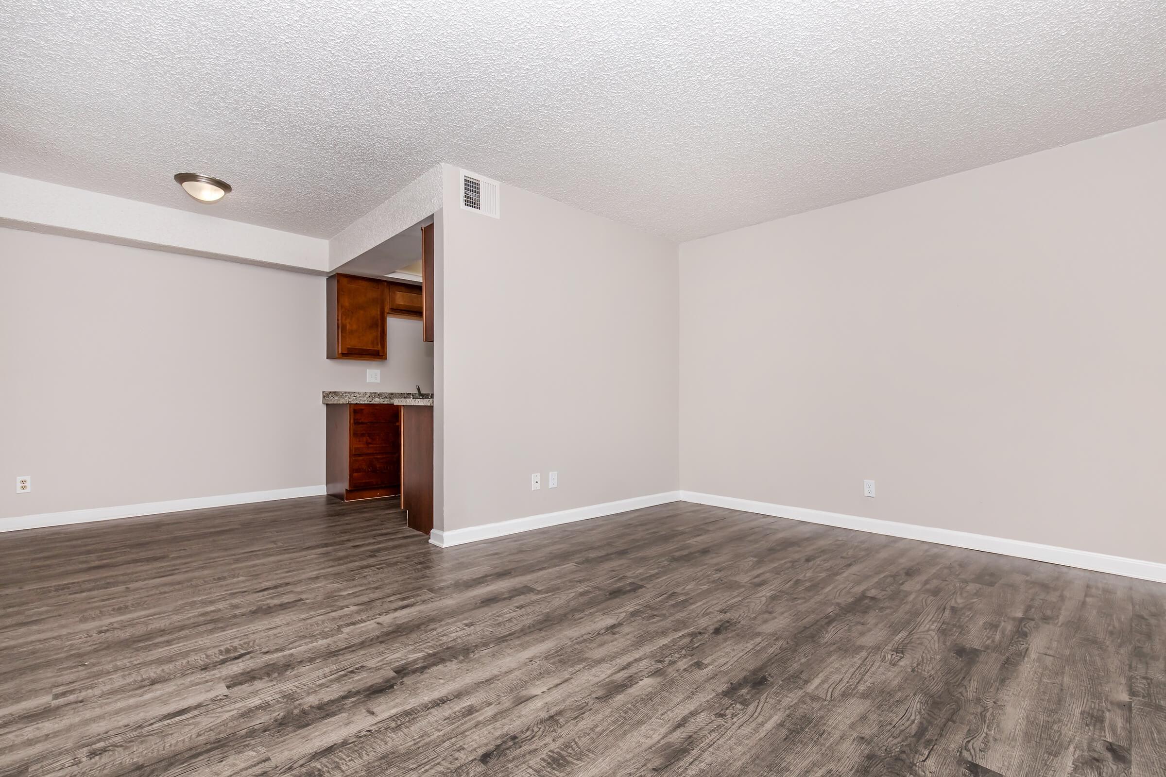 A spacious, empty room with light-colored walls and a textured ceiling. The floor is covered with dark wood-like laminate. In the background, there is an archway leading to a small kitchenette area with wooden cabinets and a countertop. The overall atmosphere is bright and open.