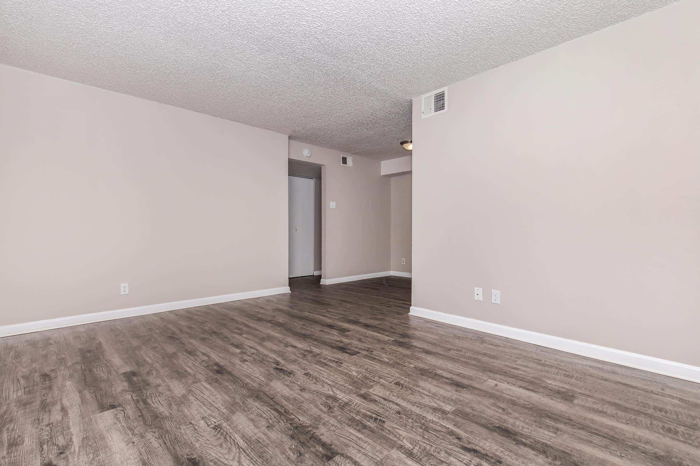 Spacious room with light-colored walls and a textured ceiling. The floor features wood-like laminate, creating a warm atmosphere. There are two doorways visible, leading to other areas, and a light fixture on the ceiling. The overall space appears bright and empty, suitable for various furniture arrangements.