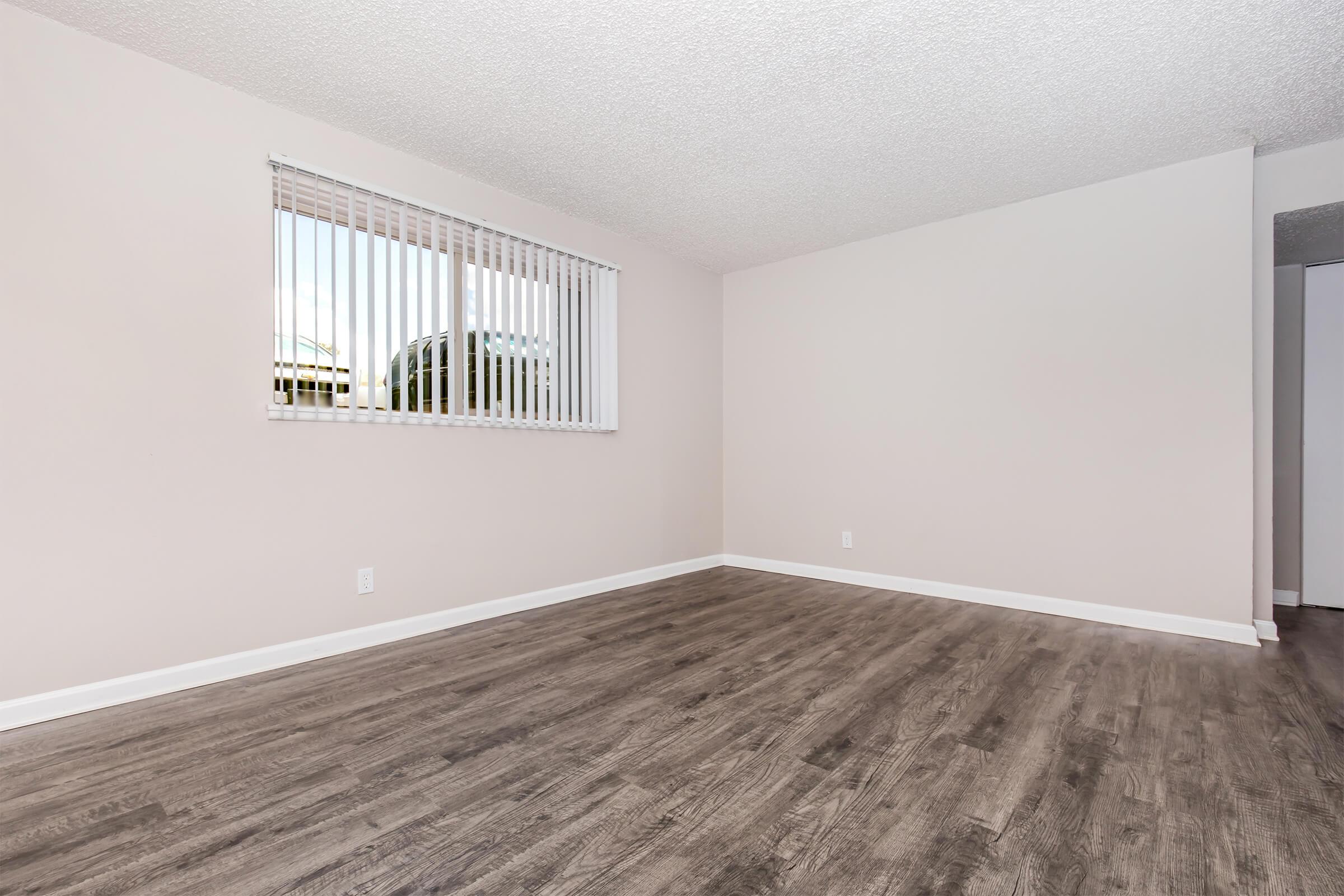 Empty room with light-colored walls and dark wooden flooring. A window with white blinds allows natural light to enter, illuminating the space. There are no furnishings or decorations, creating a clean and minimalistic atmosphere. The room features a simple aesthetic, ideal for various design possibilities.