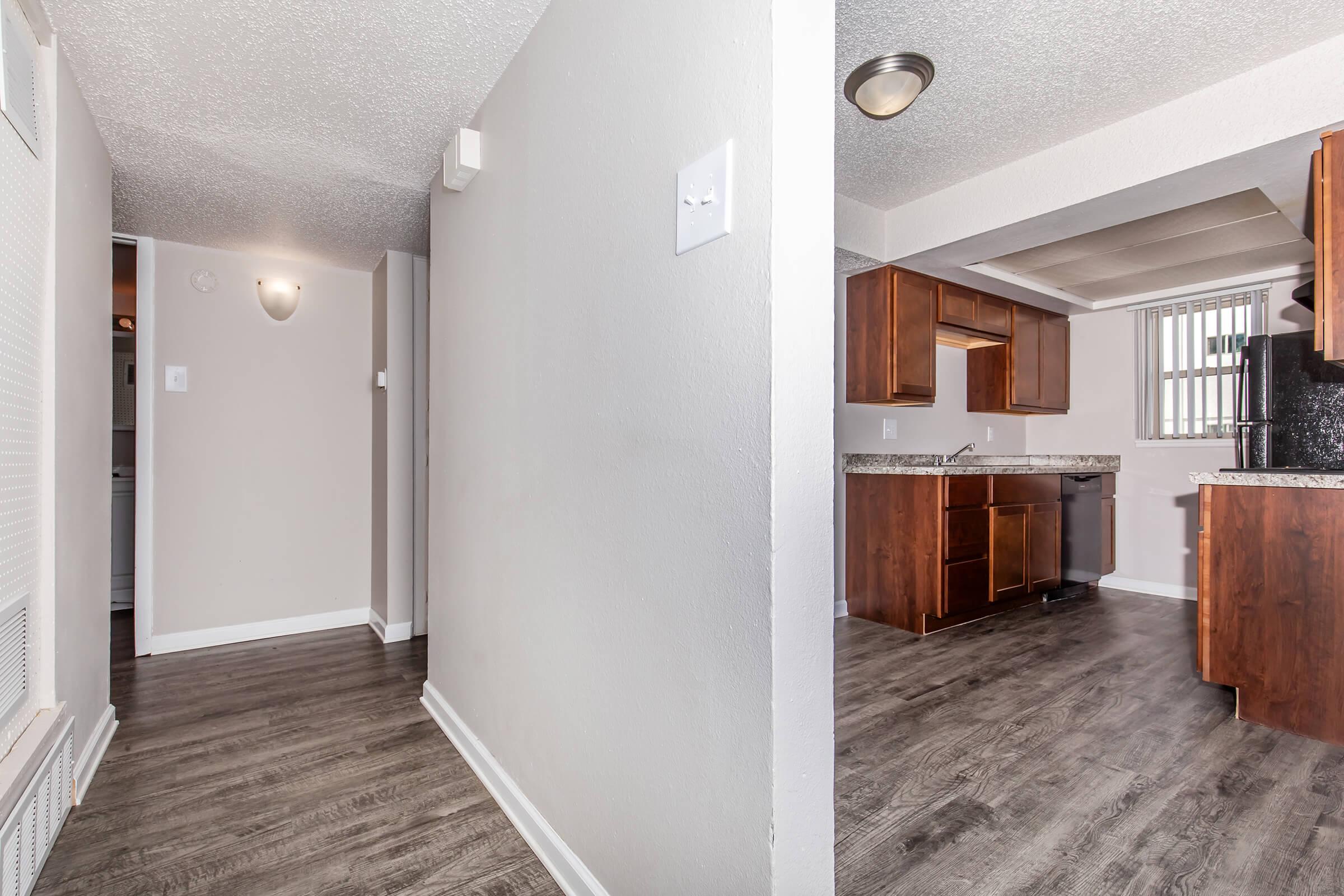 View of an interior space featuring a hallway leading to a kitchen area. The hallway has a light-colored wall and flooring, while the kitchen showcases wooden cabinets and a granite countertop. Natural light illuminates the space, creating a welcoming atmosphere.