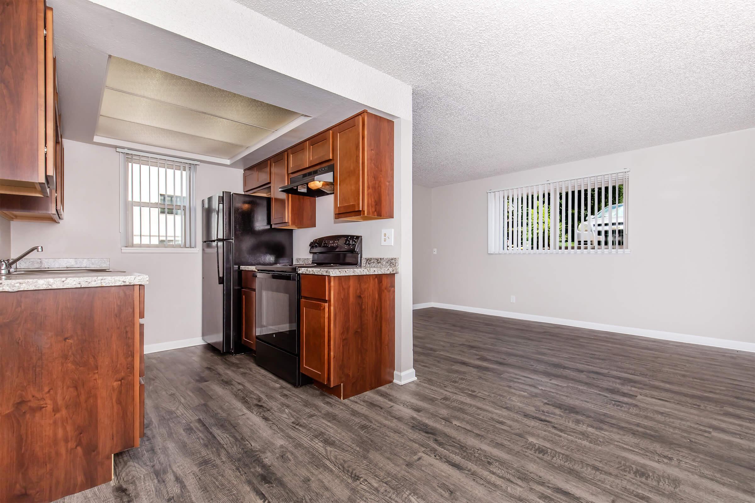 Spacious kitchen with wooden cabinets, stainless steel appliances, and granite countertops, leading into an open living area with large windows and modern flooring. Natural light fills the space, creating a bright and welcoming atmosphere.