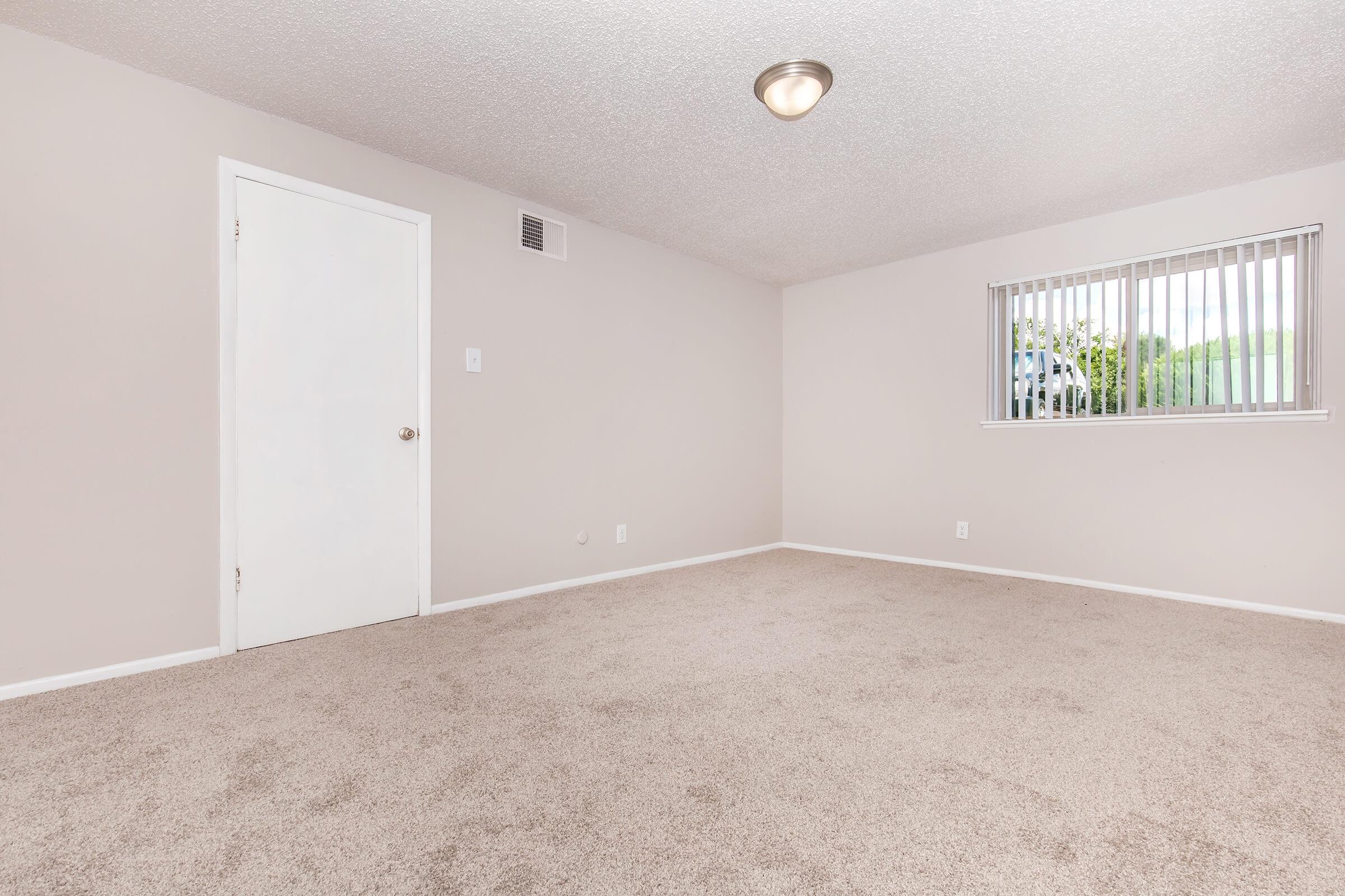 Empty room with light beige walls and carpeting. A white door leads to another space, and a window with vertical blinds allows natural light to enter. The ceiling has a simple light fixture, and there are no furnishings or decorations in the room, creating a spacious and neutral atmosphere.