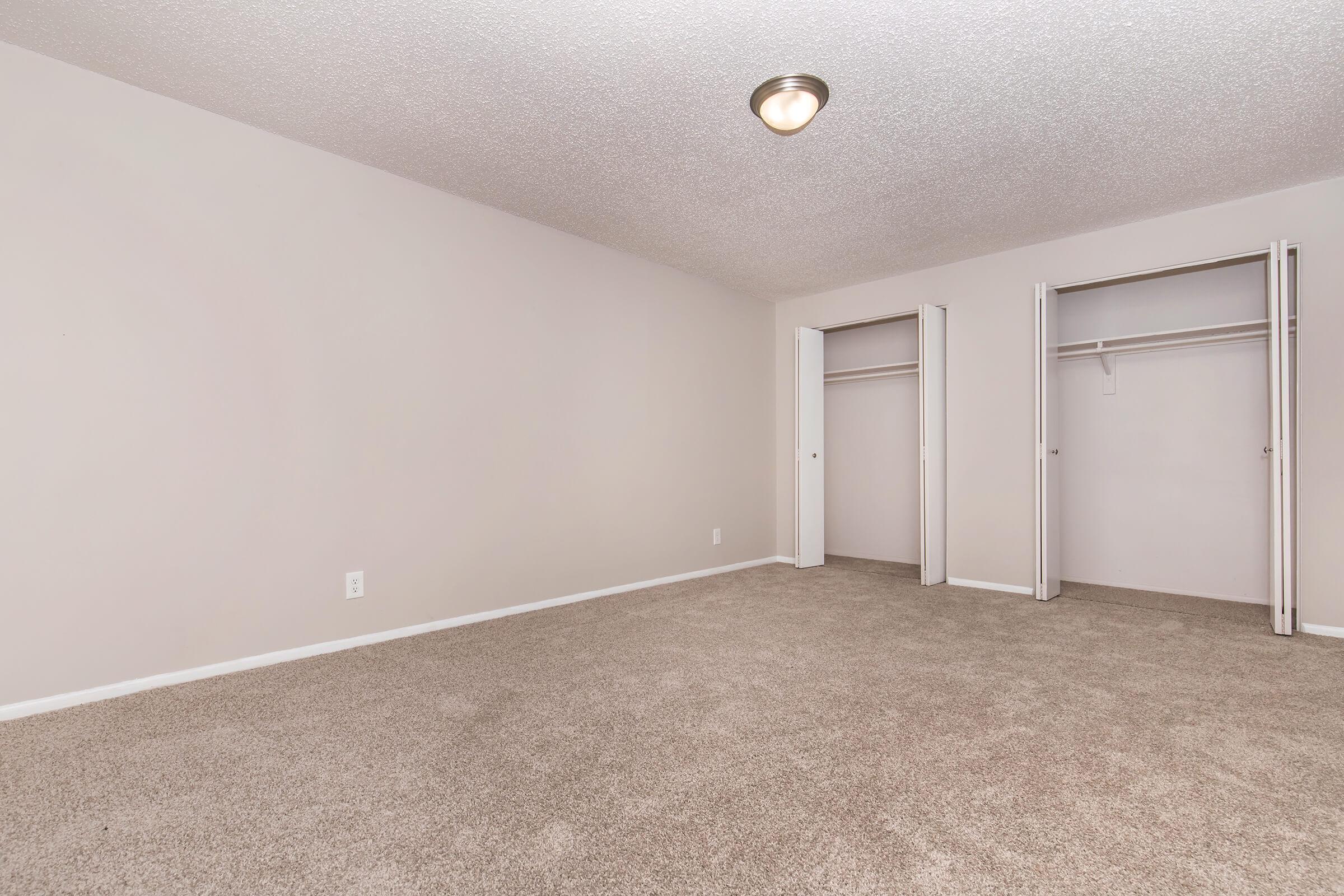 A spacious, empty bedroom with light beige walls and carpeting. The room features a ceiling light fixture and two double-door closets without doors, providing ample storage space. The overall environment is clean and unfurnished, ideal for arranging personal furniture and decor.