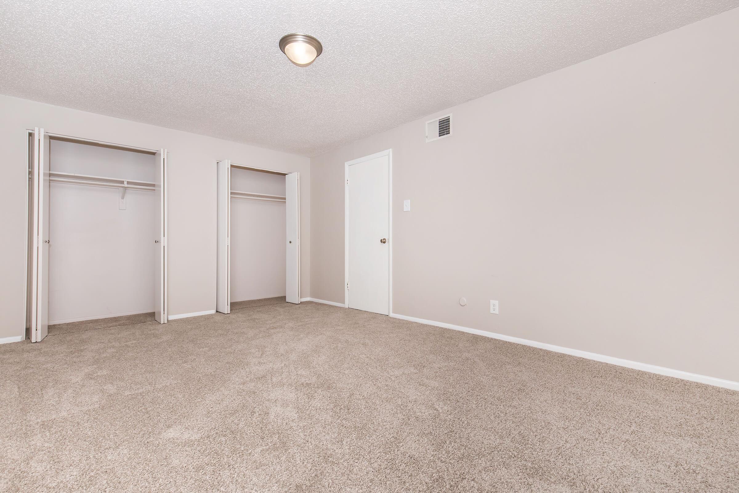An empty room with beige carpet and light gray walls. It features two open closets on the left side and a closed white door on the right. The ceiling is equipped with a simple light fixture, and there's minimal furniture or decoration, giving the space a clean and straightforward look.