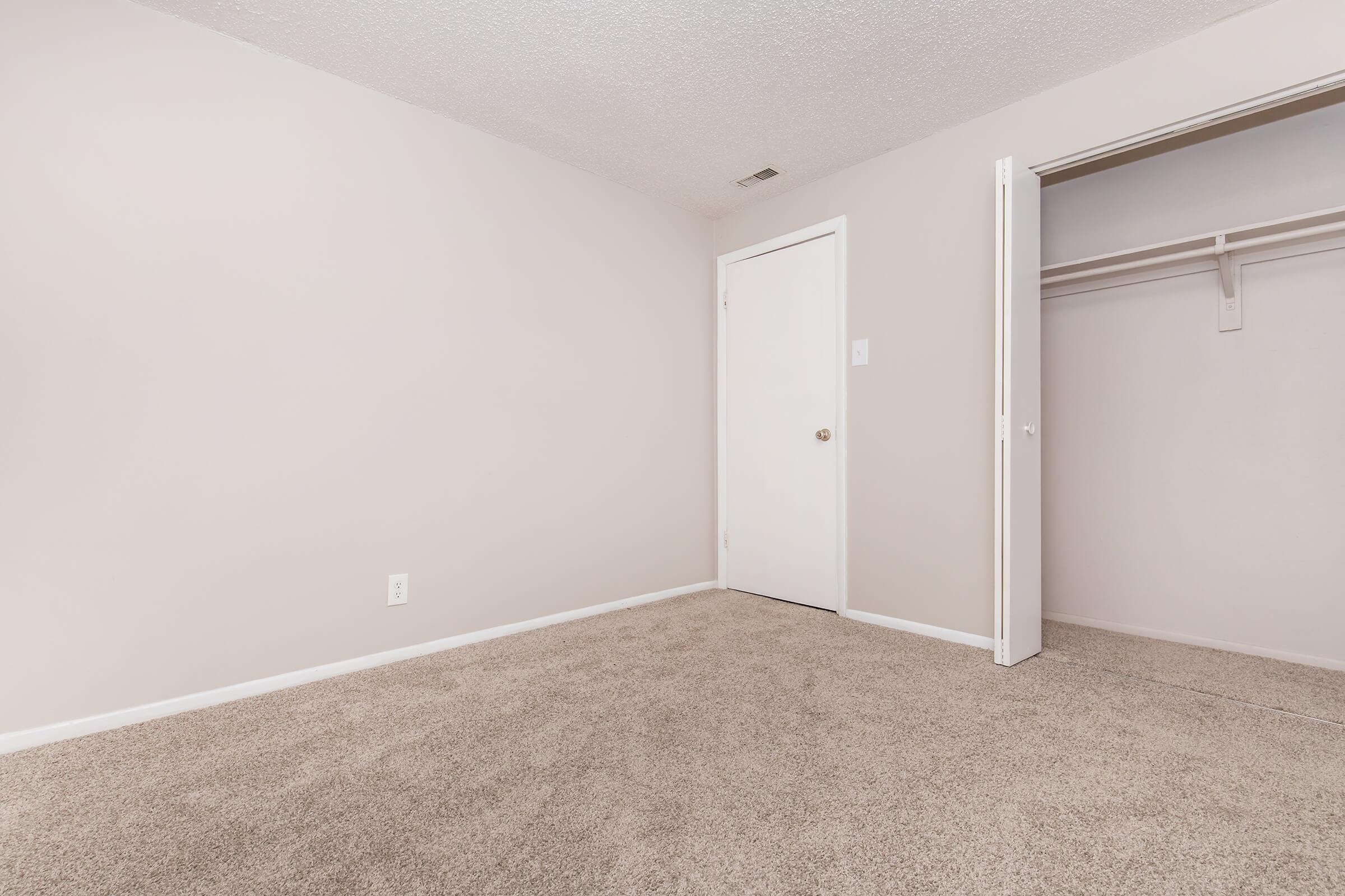 A simple, empty room with light beige walls and carpet. There is a white closet with a sliding door on the right, and a closed door on the left. Natural light may illuminate the space, creating a bright, airy atmosphere. The room is unfurnished and ready for customization.