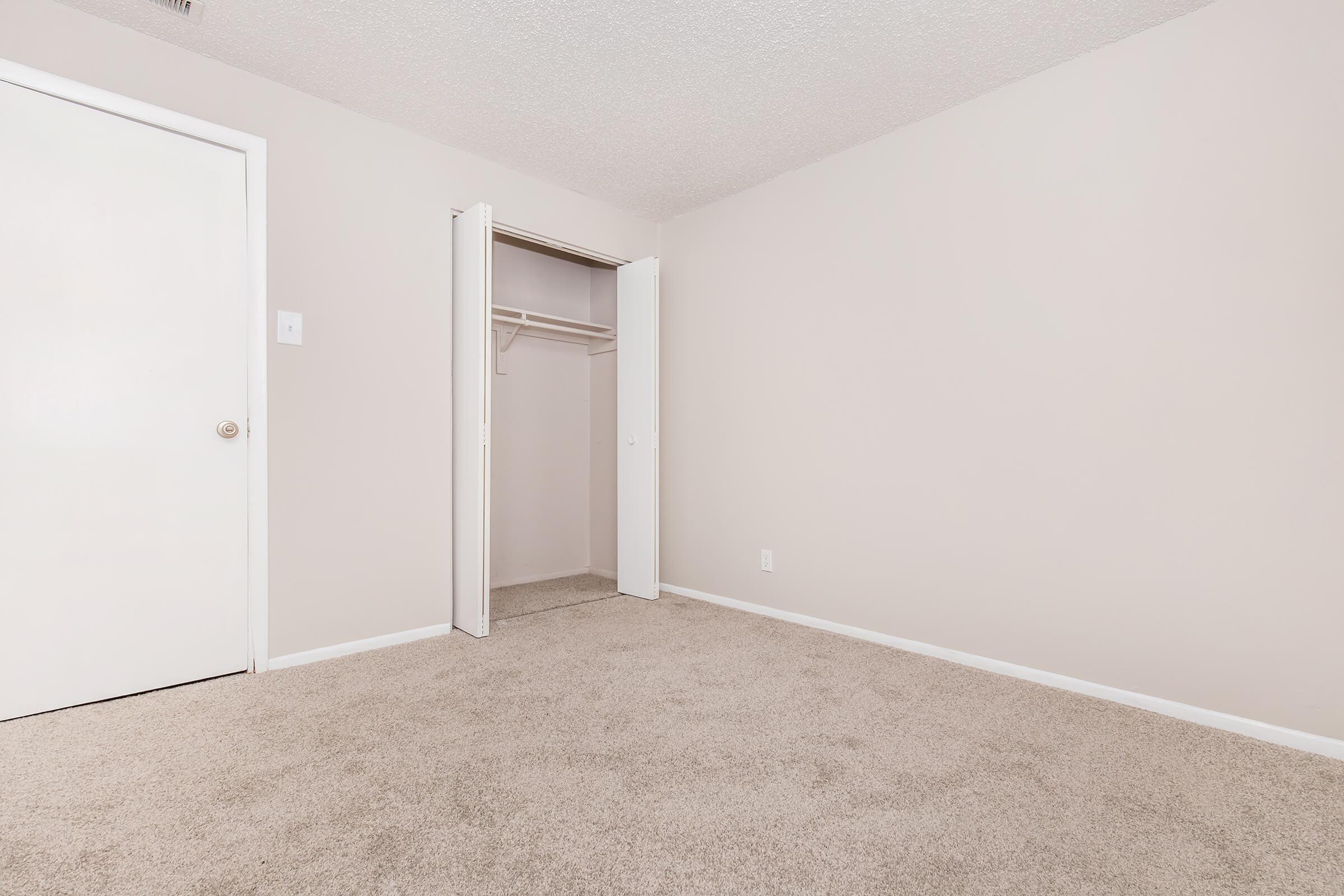 A tidy, empty room with light beige walls and carpet. There is an open closet with sliding doors on one side and a closed white door on the other. The room appears spacious and well-lit, featuring a simple, minimalistic design.