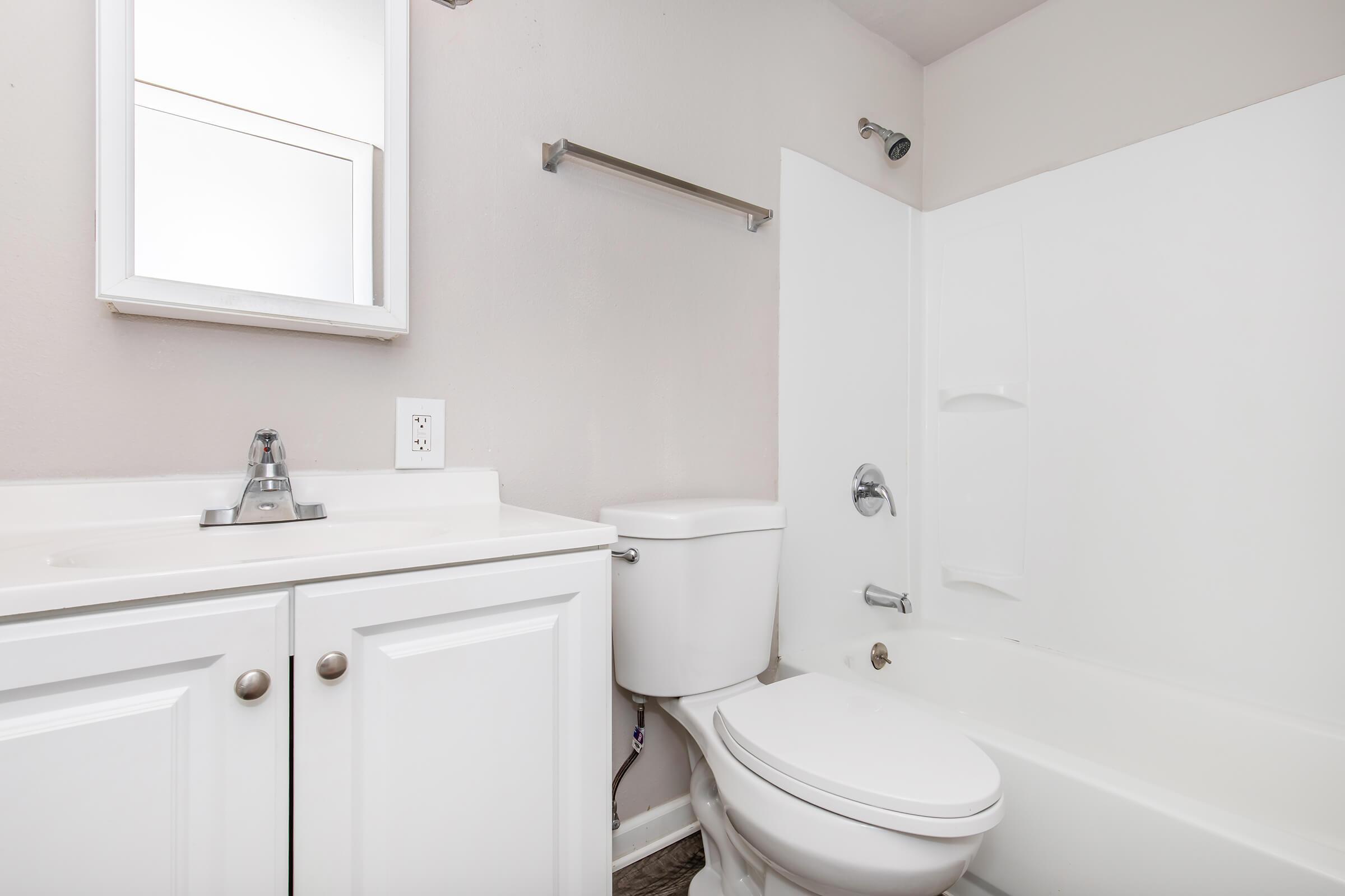 A clean, modern bathroom featuring a white bathtub with a shower, a white toilet, and a sleek vanity with a sink. There is a wall-mounted mirror above the sink and a towel bar nearby, all set against a neutral wall color. The flooring is a light wood or laminate style.