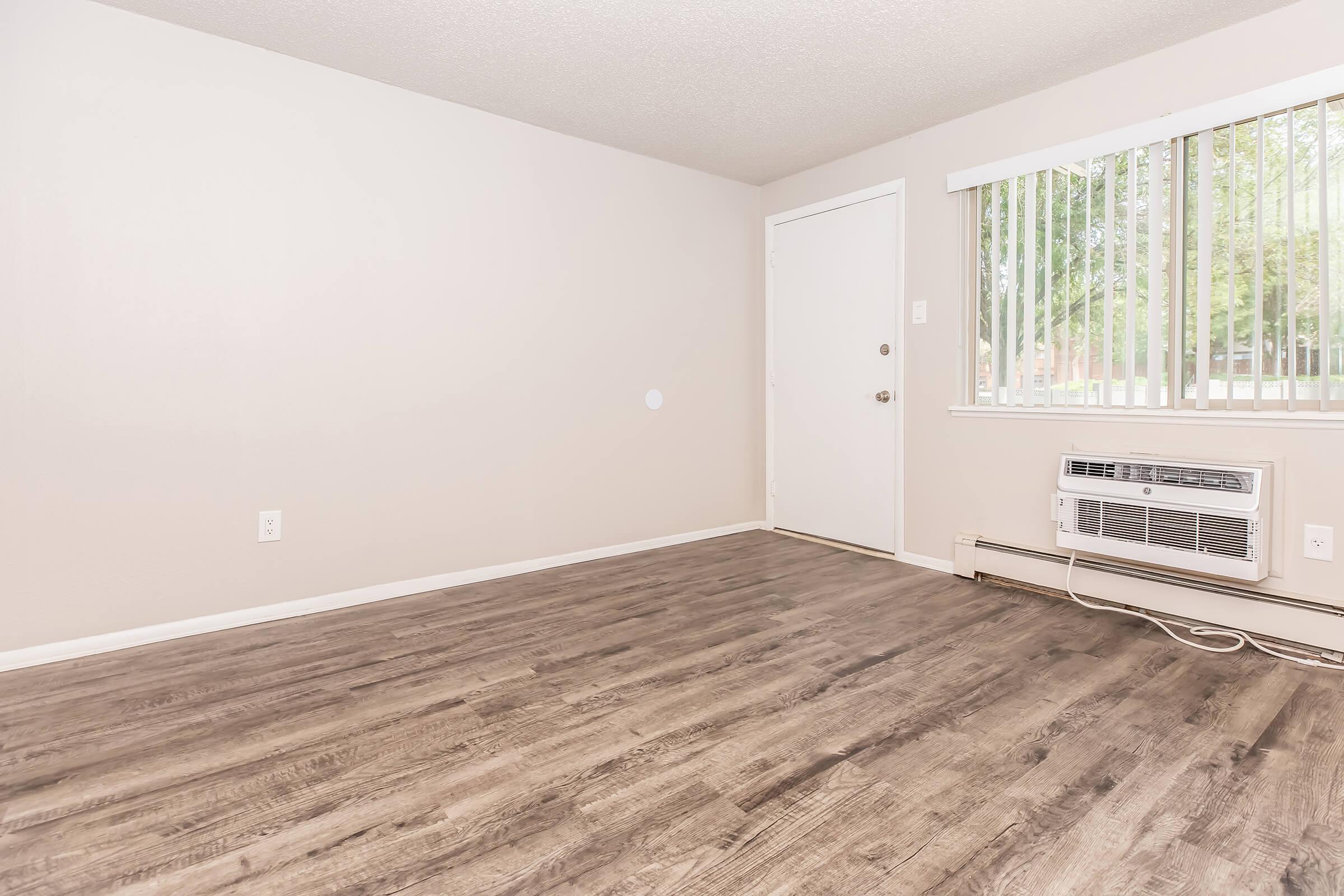 A spacious, empty room with light brown laminate flooring, beige walls, and a large window with vertical blinds. The room features an air conditioning unit on the wall and a plain door. The space is well-lit, creating a welcoming and neutral atmosphere.