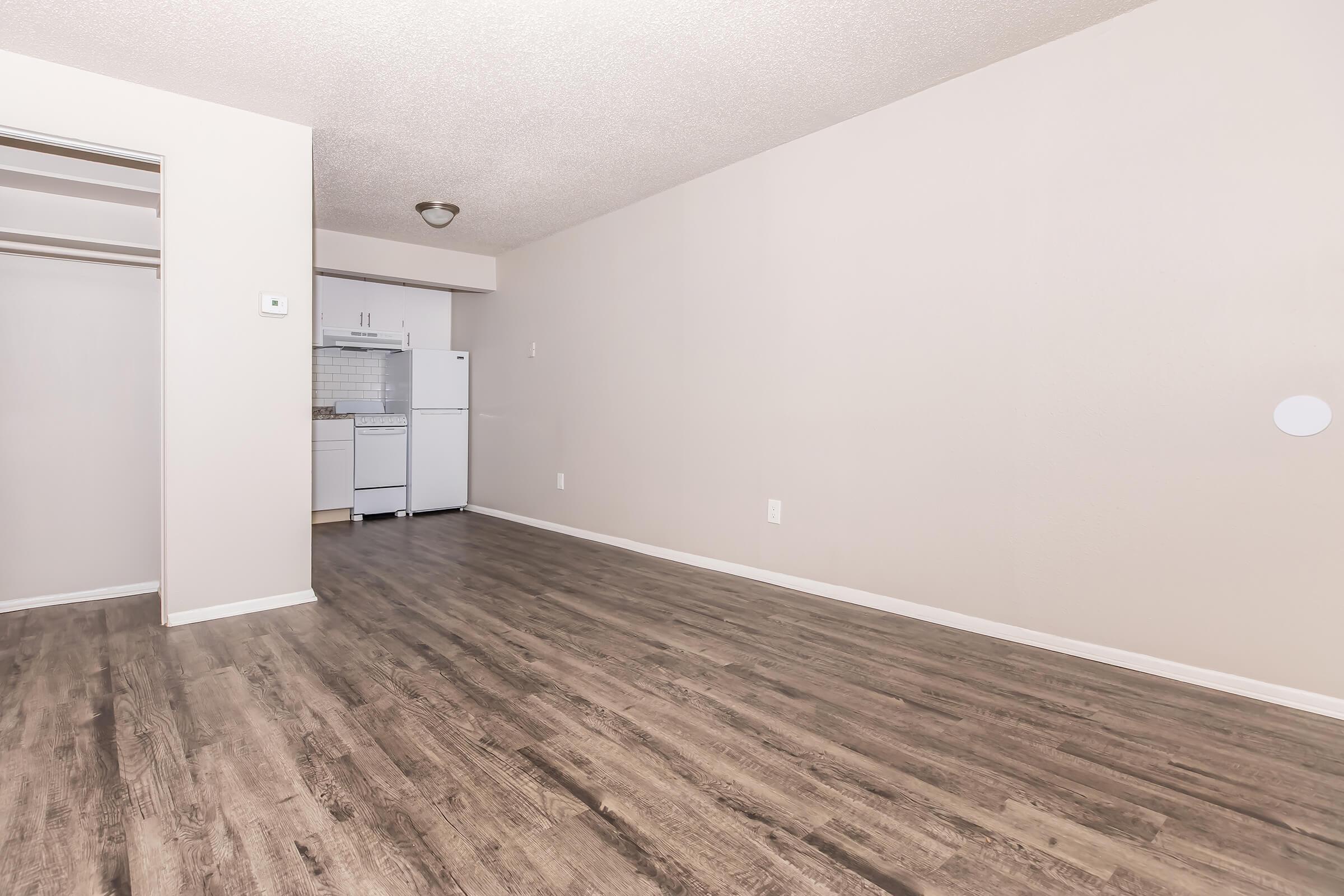 An empty room with a light-colored wall and wooden flooring. To the left, there is an open closet space. In the background, a kitchen is visible with white appliances, including a refrigerator. The area is brightly lit, creating an open and spacious feel.