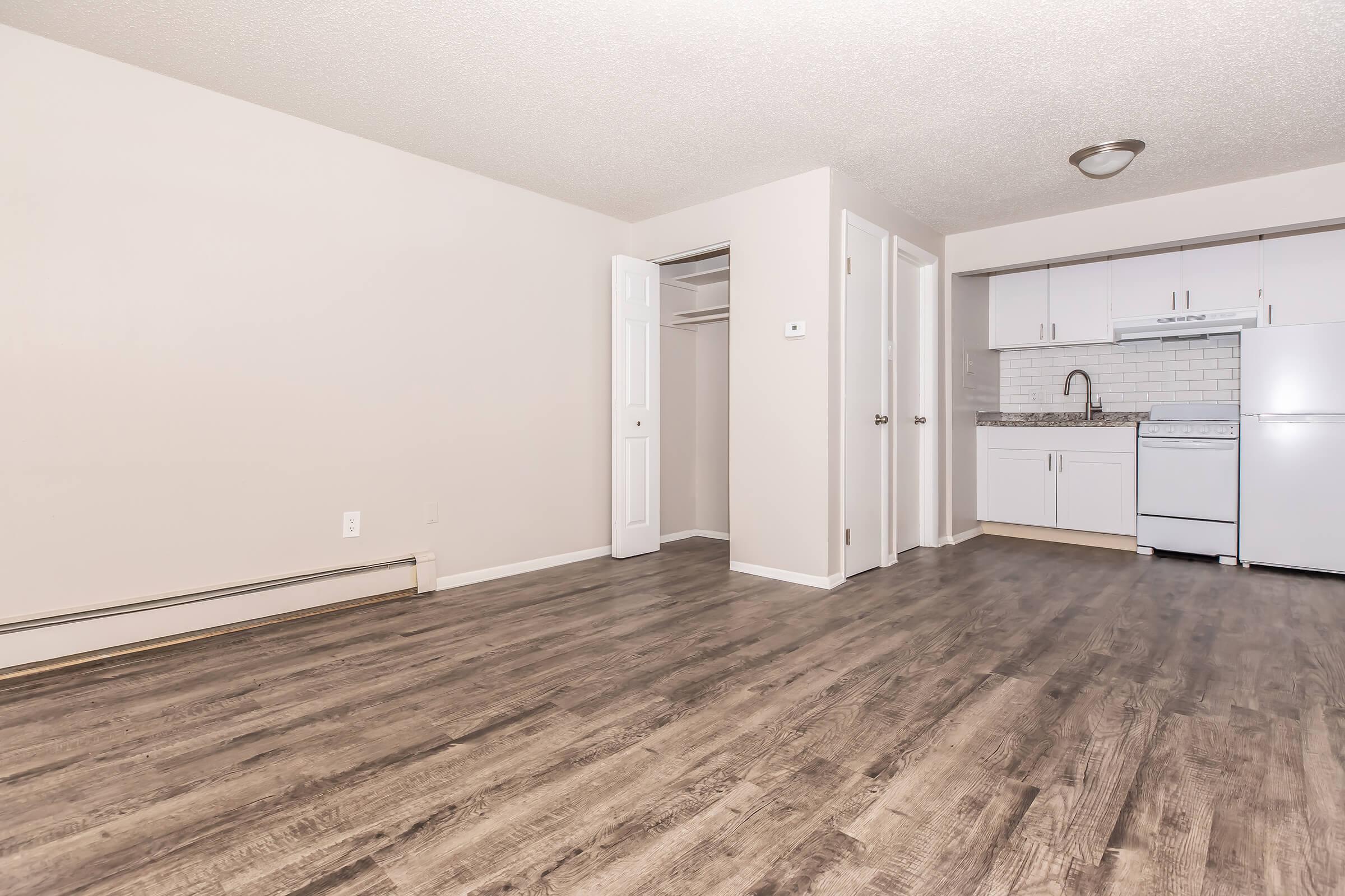 A spacious, light-colored living area featuring wood-like flooring. On one side, there is a small kitchen with white cabinets and appliances, including a refrigerator. There is also a closet with white sliding doors in the corner. The walls are painted a neutral shade.
