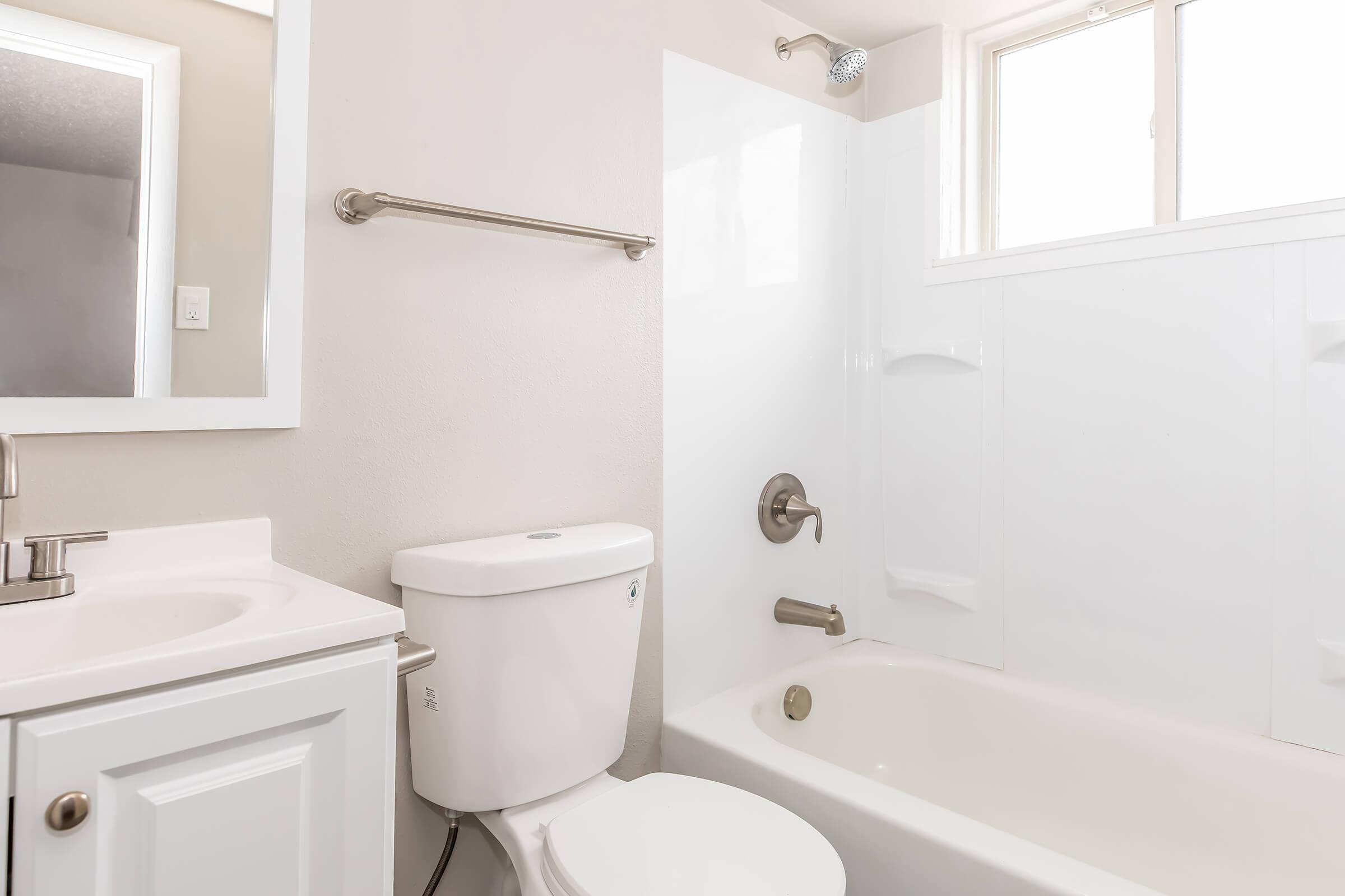 A clean, modern bathroom featuring a white bathtub with a showerhead, a toilet, a sink with a mirror above, and light-colored walls. Natural light enters through a window, enhancing the bright and airy atmosphere.