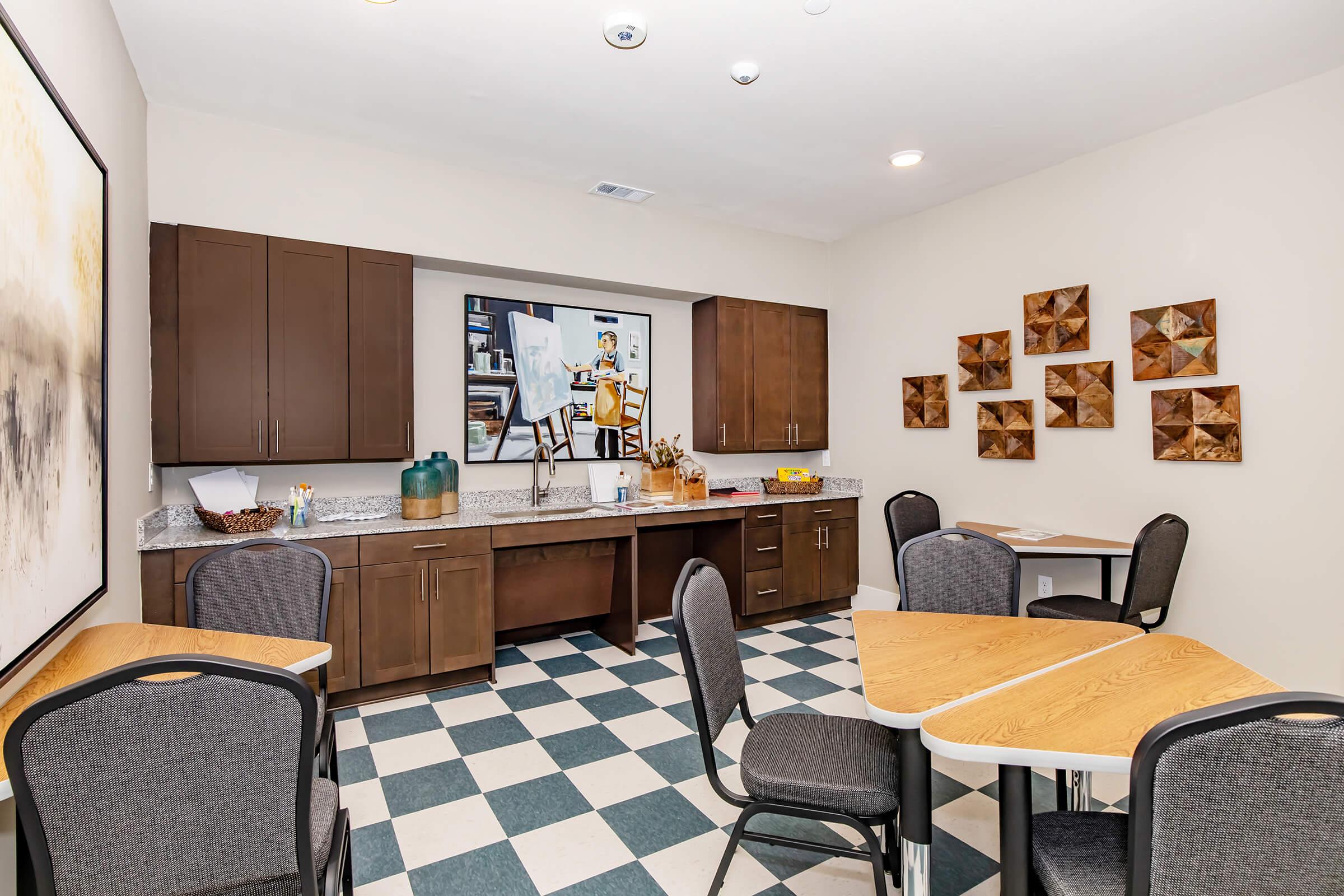 A well-lit common area features a countertop with a large painting above it, decorated with art supplies and snacks. The flooring has a black and white checkerboard pattern. There are several tables with chairs arranged neatly, and decorative wooden wall art adds a touch of warmth to the space.
