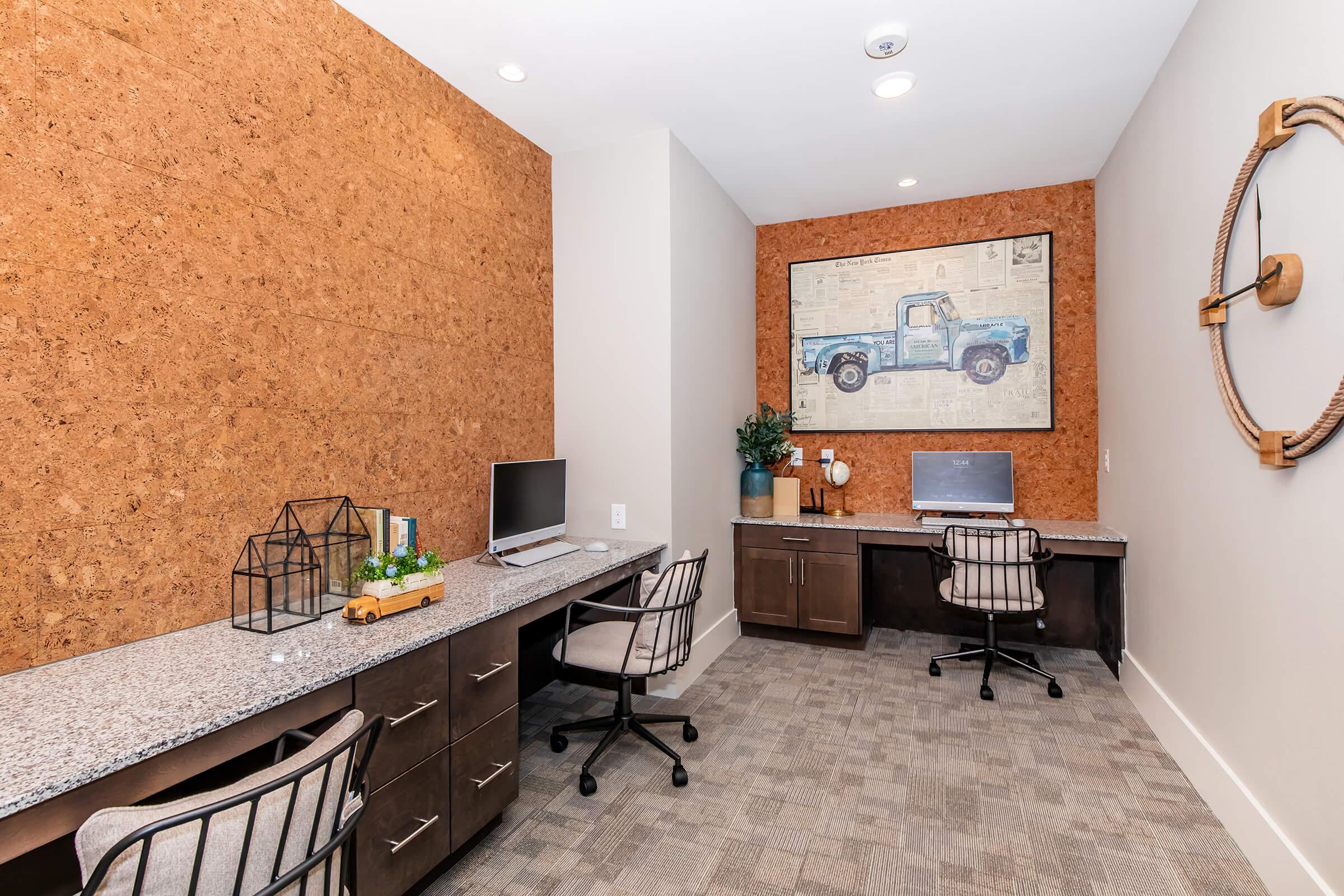 A modern office space featuring two workstations with computers, grey granite desks, and ergonomic chairs. The walls include a textured cork panel and a large framed artwork of a blue vintage truck. A small green plant and decorative items are arranged on a desk, adding a touch of style.