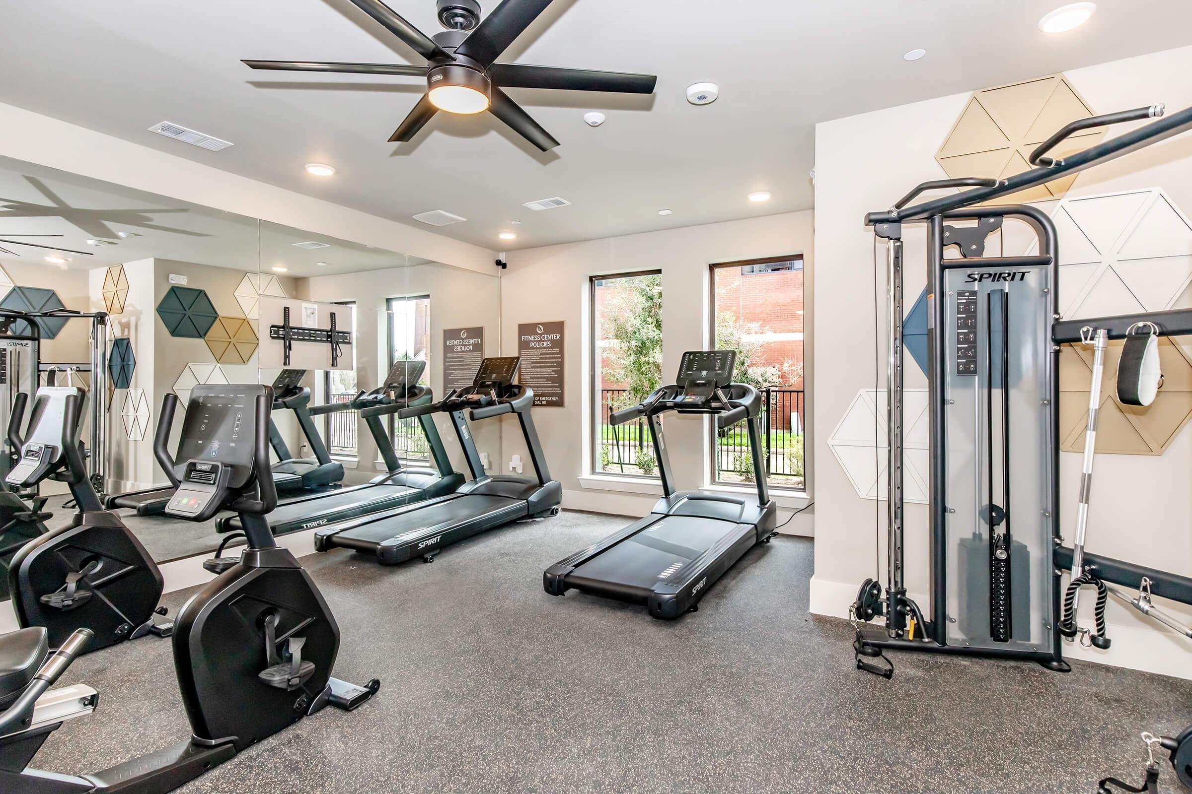 A modern gym interior featuring various exercise equipment including treadmills, stationary bikes, and a weight machine. Large windows allow natural light to fill the space, and the walls are designed with geometric patterns. The flooring is dark and the ceiling has a fan for ventilation.