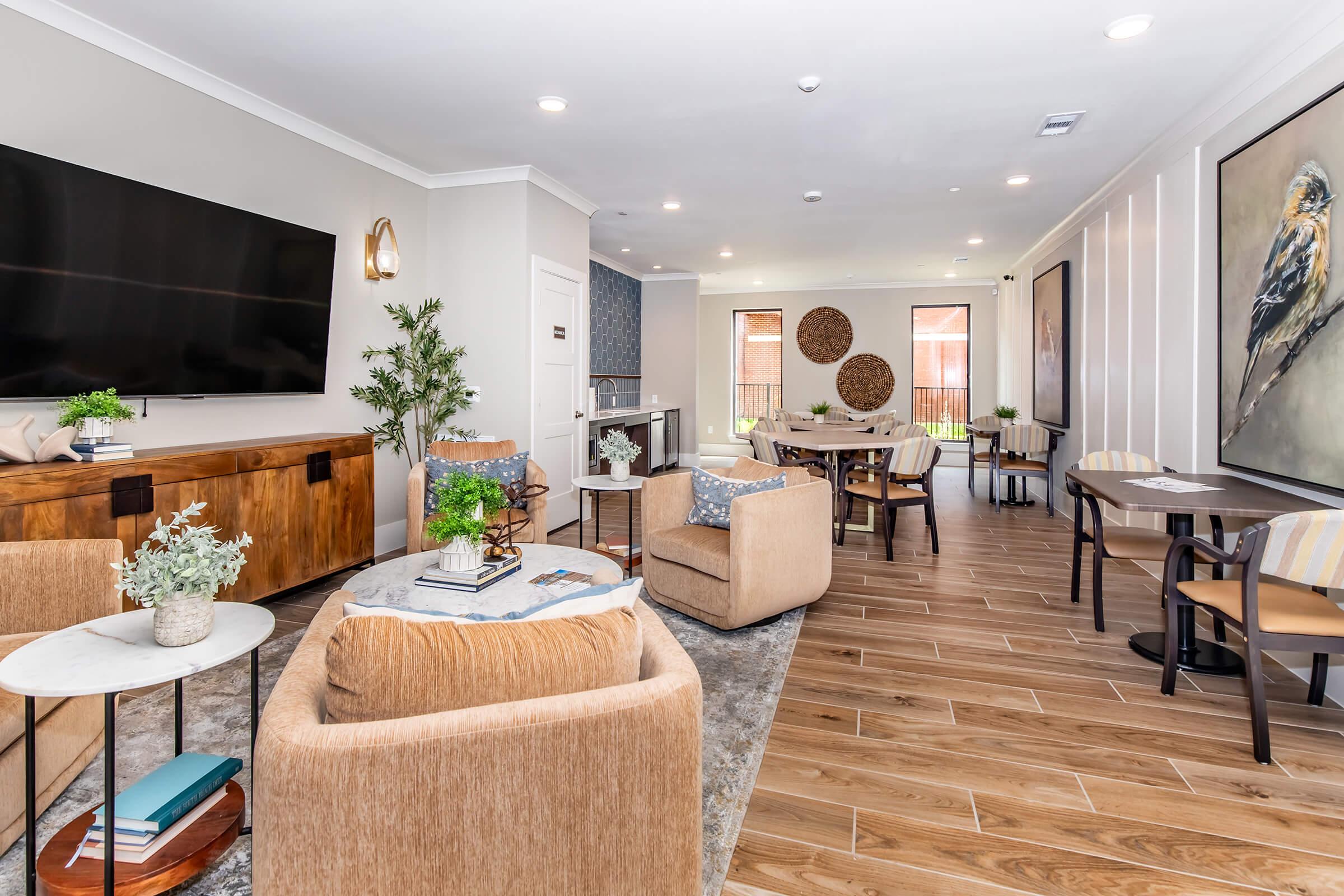 A stylish lounge area with comfortable seating, including plush armchairs and small tables. There's a large television on one wall, decorative plants, and modern artwork. Natural light filters through large windows, creating a welcoming atmosphere. The floor is wooden, enhancing the contemporary design.