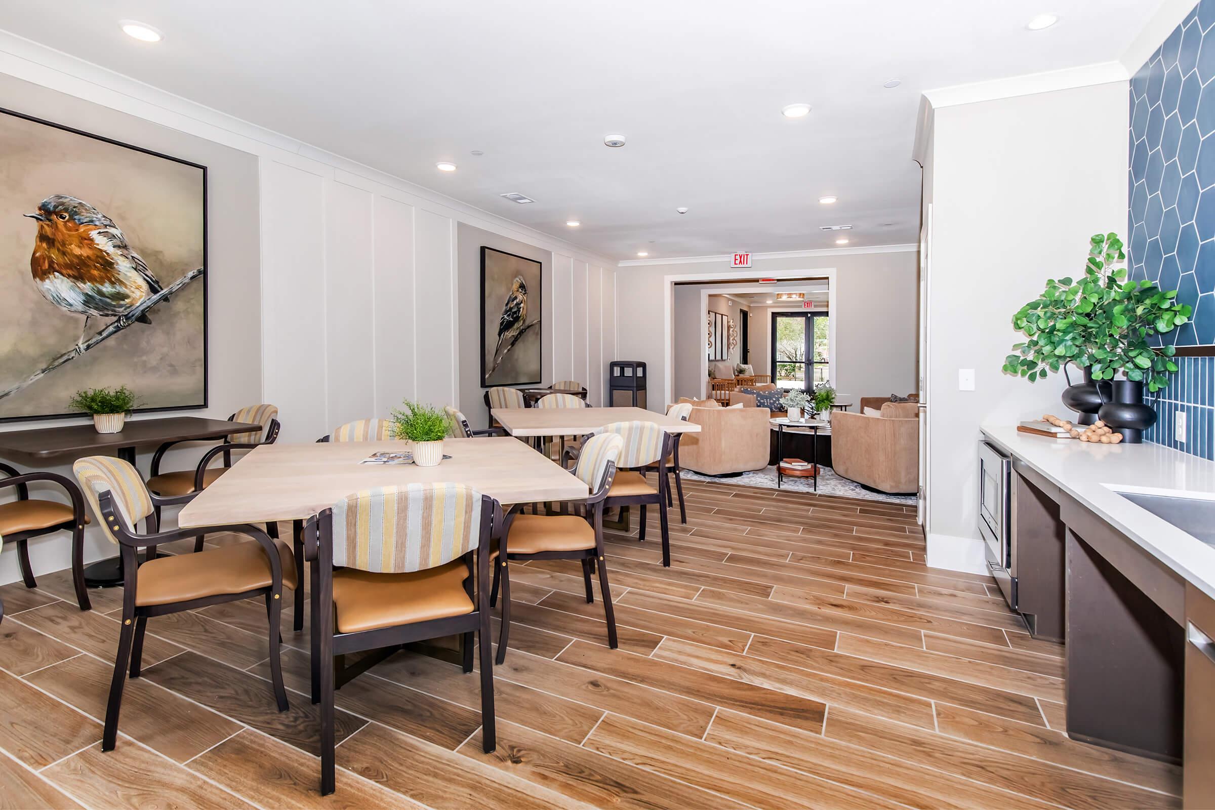 A modern, well-lit communal room featuring wooden flooring, tables with chairs, and a decorative plant on the table. Two large bird paintings adorn the walls, and a cozy seating area is visible in the background, enhancing the inviting atmosphere of the space.