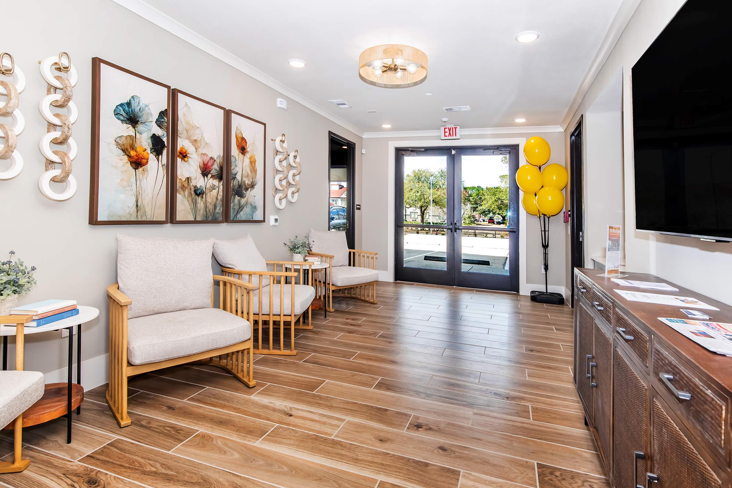 A bright waiting area featuring two light-colored armchairs and a wooden side table. On the walls, there are three framed artworks of flowers. A large flat-screen TV is mounted on one wall, and near the entrance, yellow balloons are displayed. The floor is made of wooden tiles, contributing to a welcoming atmosphere.